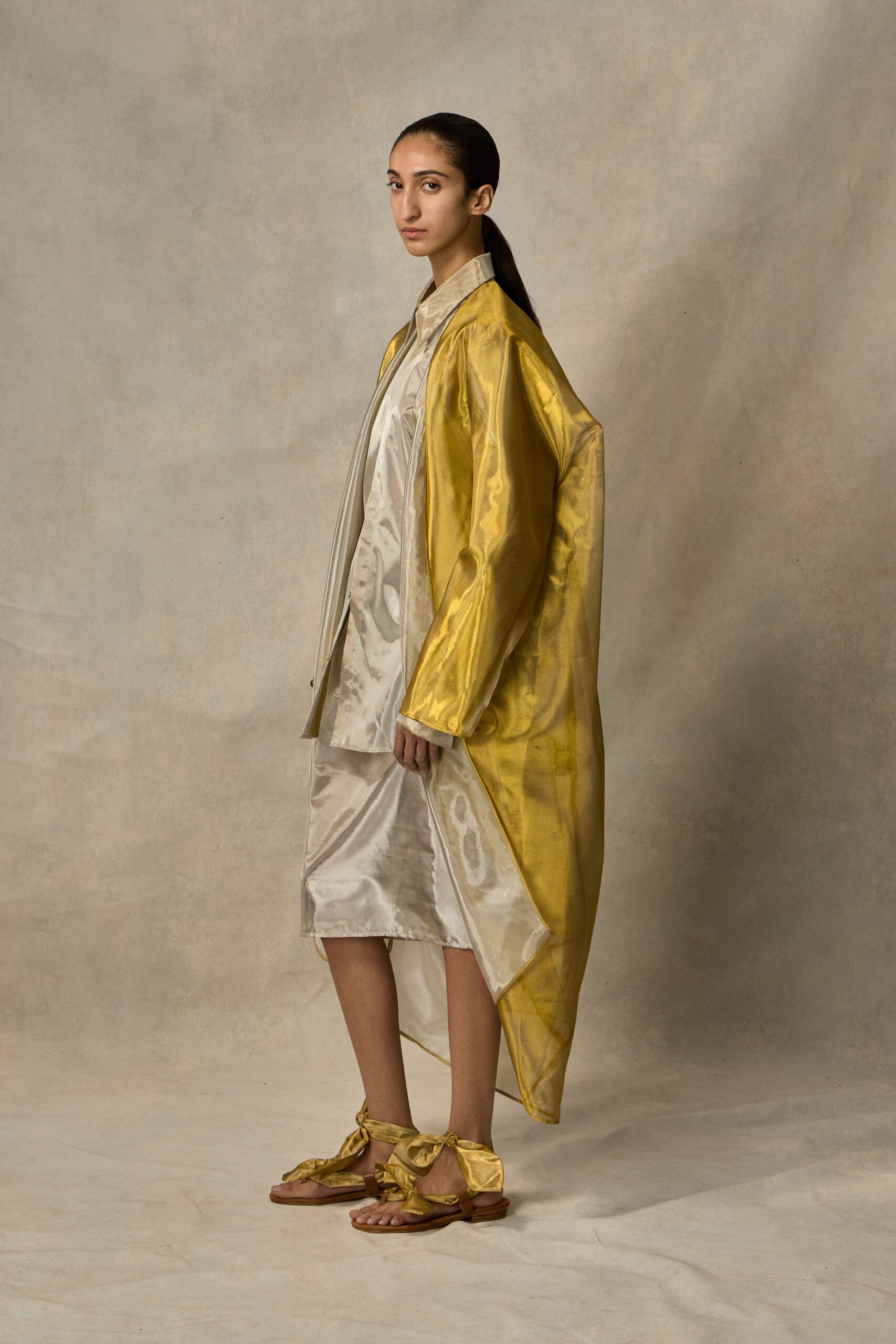 Ochre lamé coat hanging open, one side falling longer than the other. Pearl satin shirt with a soft pointed collar and loose placket. Matching satin skirt, wrinkling into watery highlights at the knee. Flat sandals tied with oversized metallic bows at the ankles. Crossing a museum courtyard just before the rain starts.