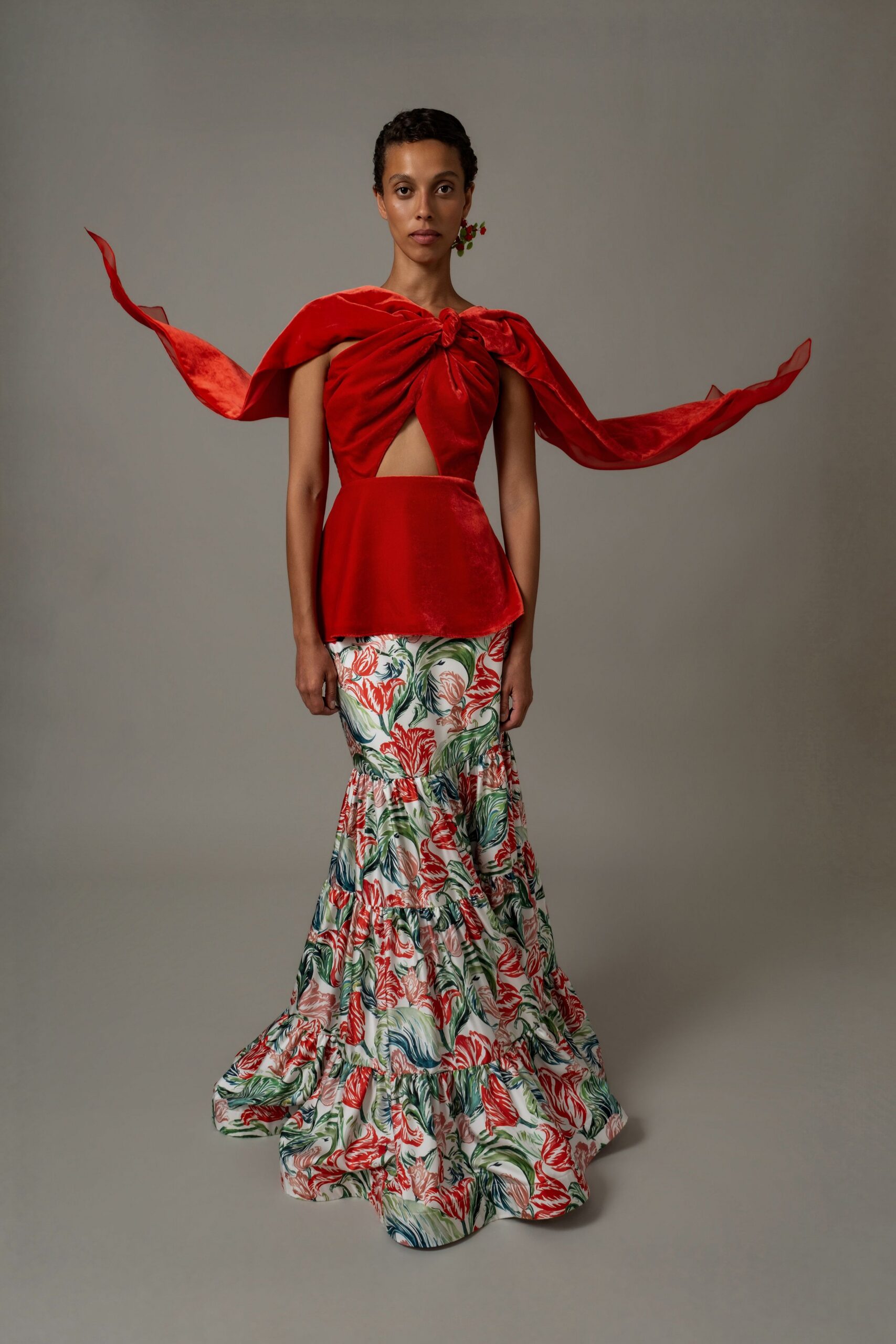 Scarlet velvet top knotted at the chest, chiffon streamers lifting from the shoulders. Tiered mermaid skirt in painterly coral and seaweed print, hem pooling wide. Small red berry cluster at one ear. Garden wedding, wind coming off the water before the plates are cleared.