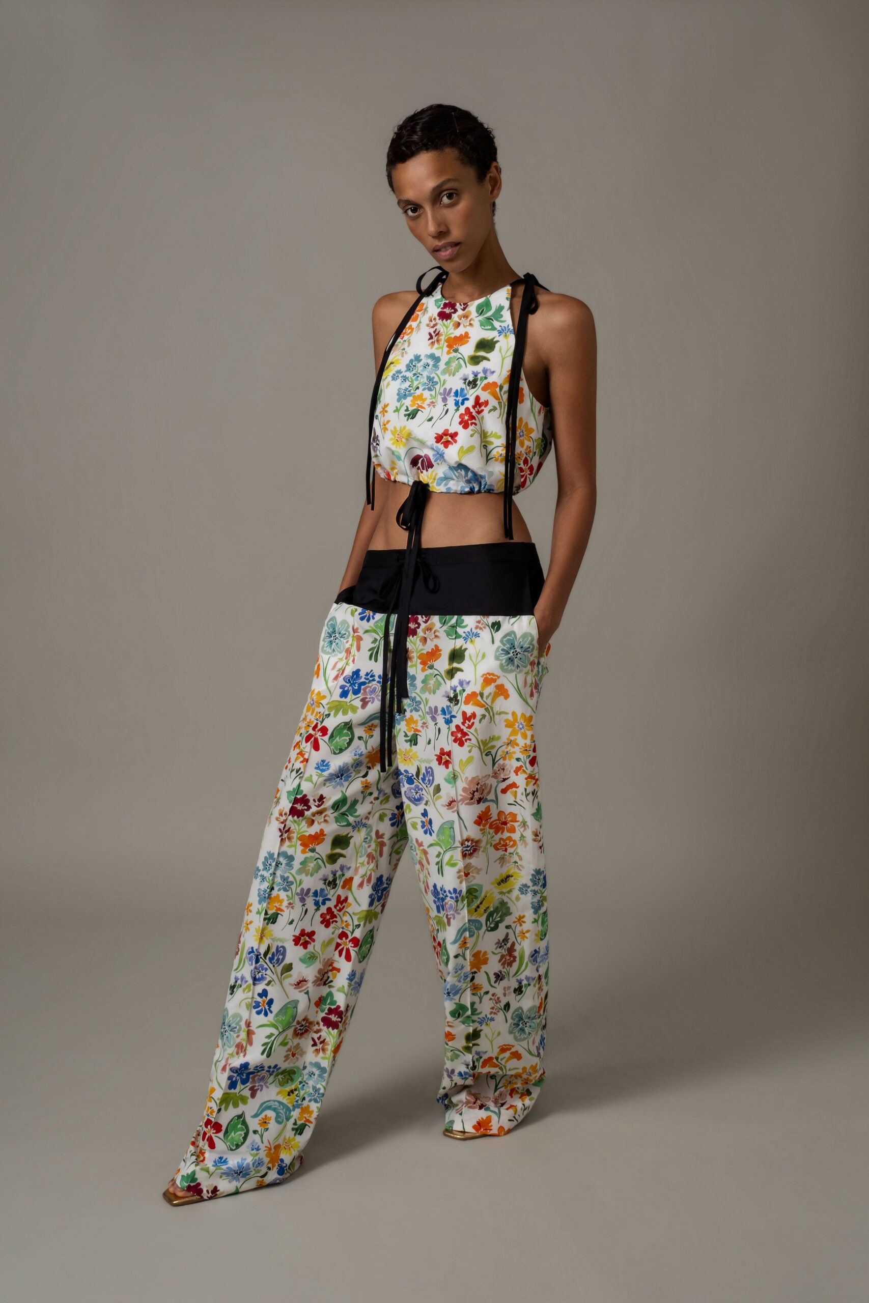 White floral halter top ruched under the bust with black ties. Wide white floral trousers with a deep black waistband and slant pockets. Square-toe metallic sandals peeking out under the pooling hem. Walking back from a garden wedding after the grass has flattened.