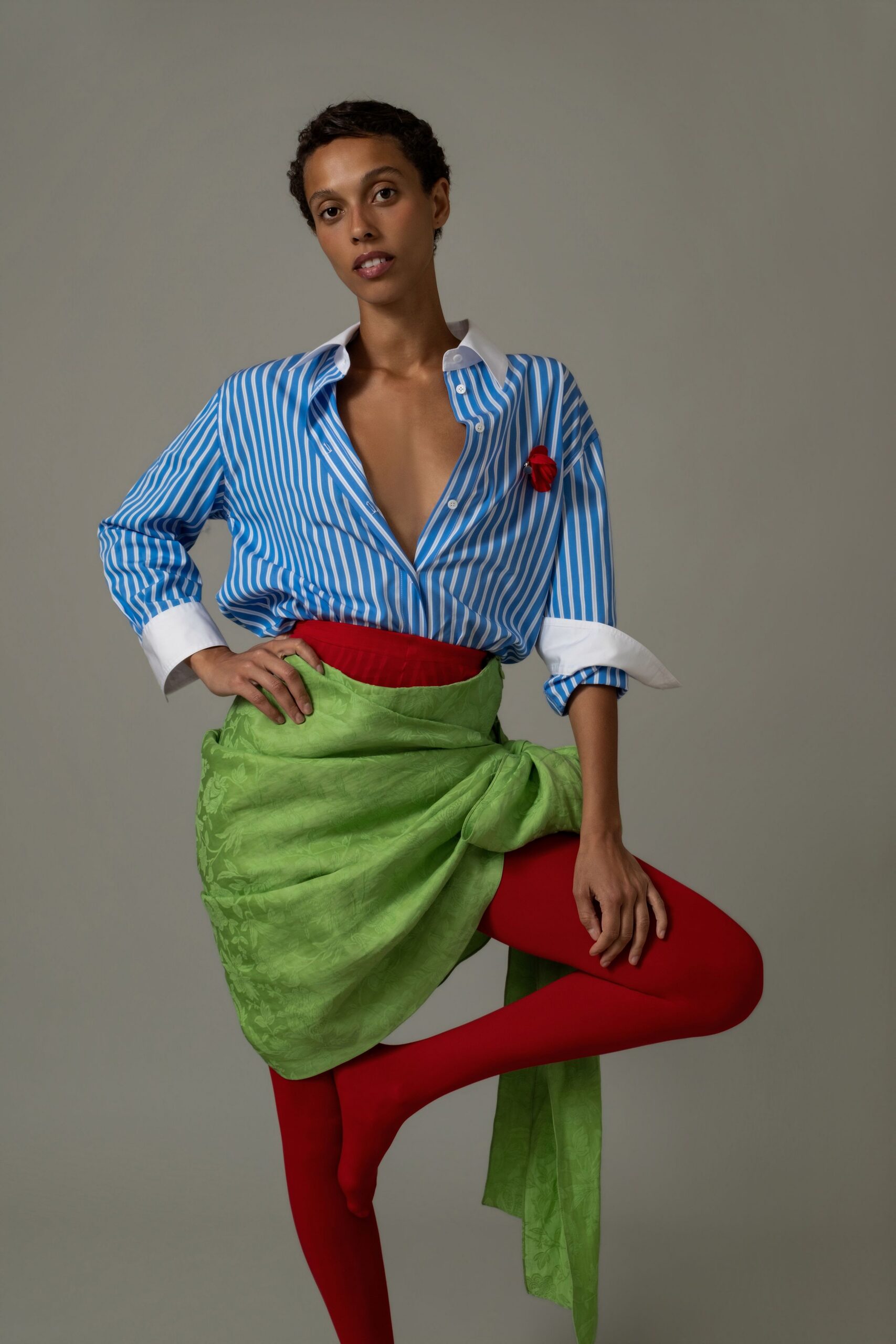 Blue-and-white banker-striped shirt with a white spread collar and turned cuffs. Small red rosette pinned at the chest. Acid green floral jacquard wrap knotted into a thick side twist. Opaque scarlet tights, unbroken from waist to pointed toe. Walking into a winter garden party with the table linens still being laid.