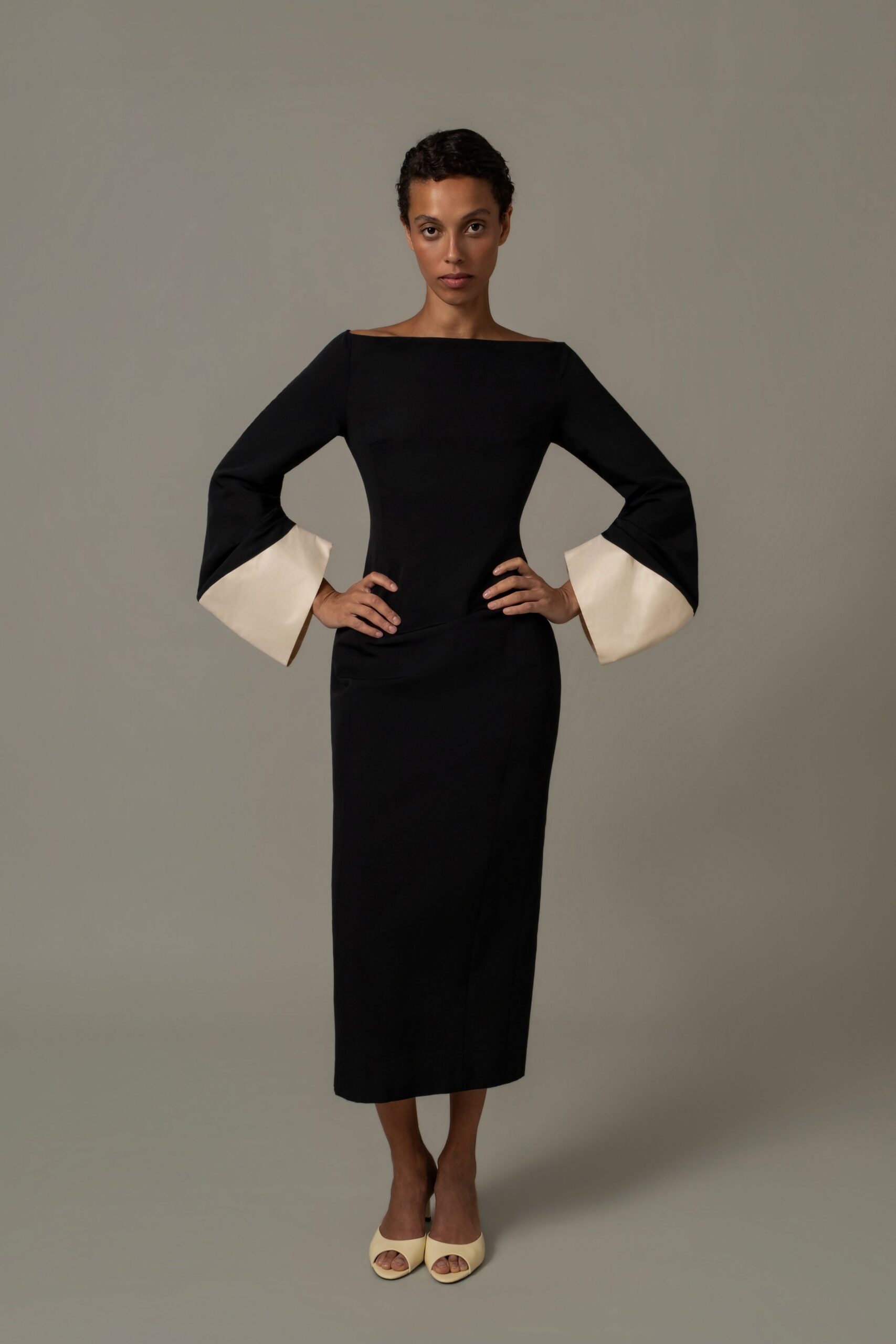 Black column dress with a straight bateau neck and ivory bell cuffs. Cream peep-toe mules with a rounded vamp. Museum benefit at noon, standing near the marble staircase before speeches.
