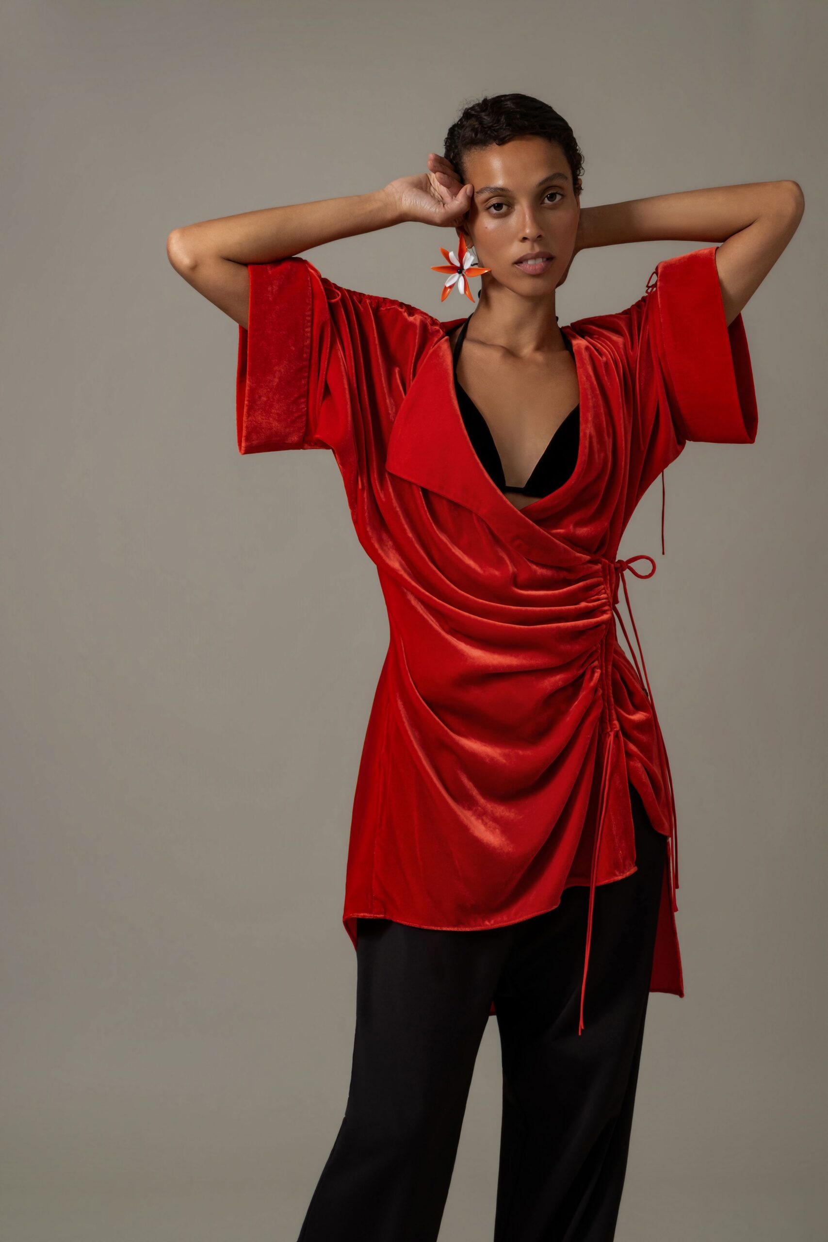 Oversized pinwheel earring in orange and white enamel petals. Red satin wrap top, ruched tight with dangling side cords. Black triangle bralette cutting sharply through the plunging neckline. Black fluid trousers with a loose straight fall. For drinks on a hotel terrace while the awnings snap in sea wind.
