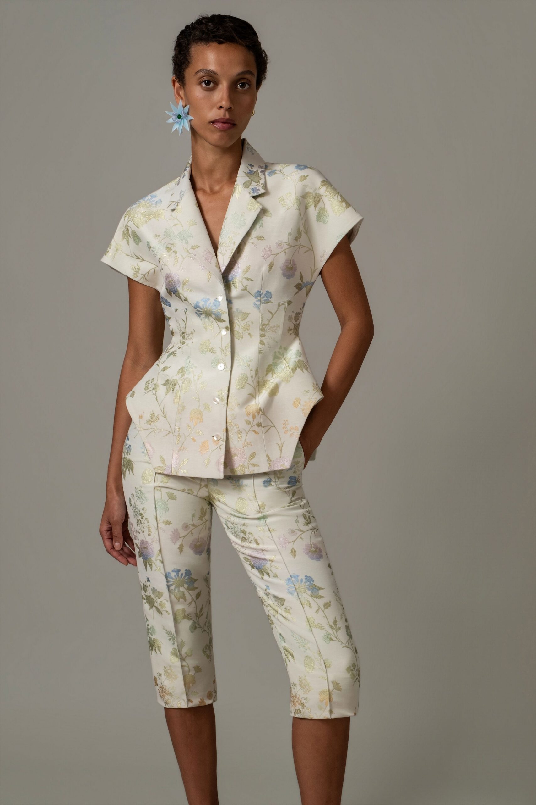 Pale blue starburst flower earring with a dark bead center. Cream floral brocade jacket, cap sleeves and peplum hips kicked outward. Matching knee-length tailored shorts, pressed front seams running arrow-straight. Garden wedding reception, cake plates cleared and the light turning silver.