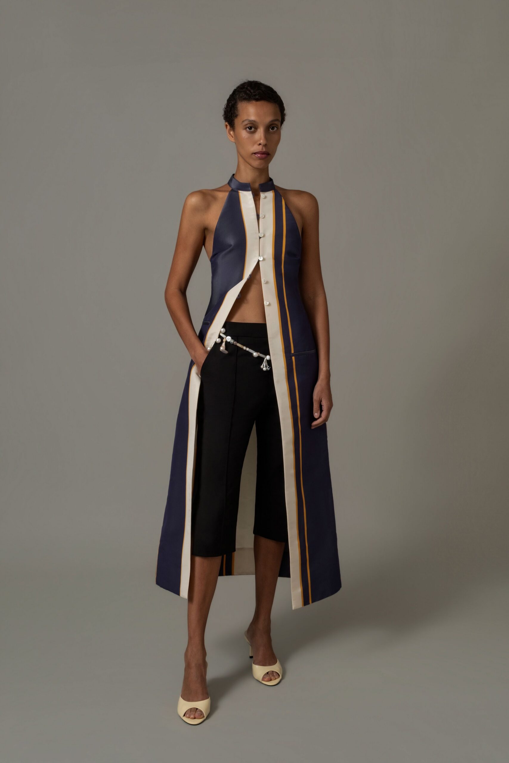 Ink halter coatdress with cream placket, split front and mustard pinstripes. Black cropped trousers with a pearl swag hooked low at the waistband. Butter peep-toe mules. For drifting through a hotel lobby after the conference badges come off.