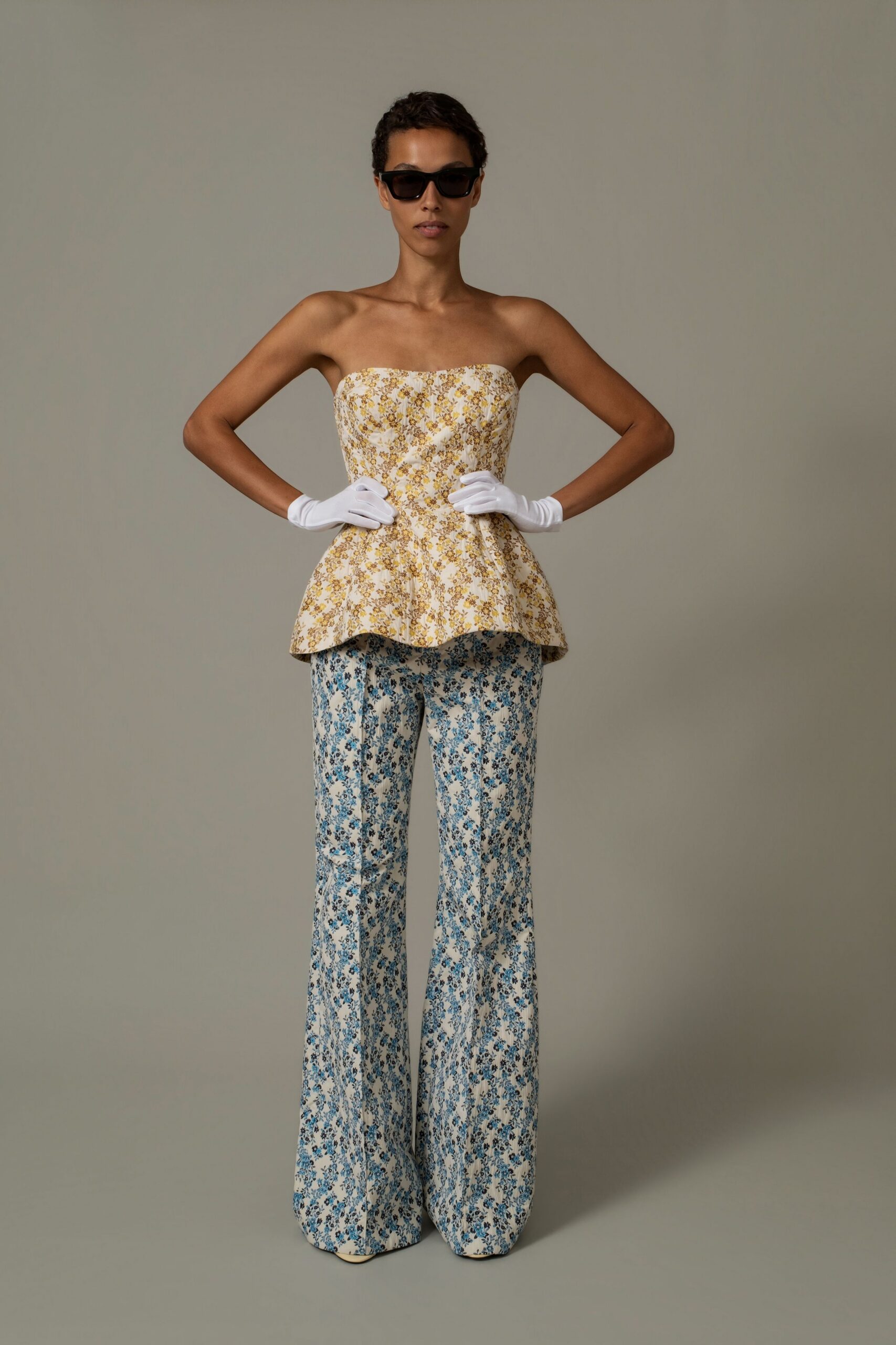 Black rectangular sunglasses with thick glossy frames. Strapless cream bustier in yellow floral brocade, peplum hem kicked out. Short white gloves. Blue floral brocade trousers falling wide enough to cover the shoes. Garden wedding reception, someone has dragged the good chairs onto the lawn.