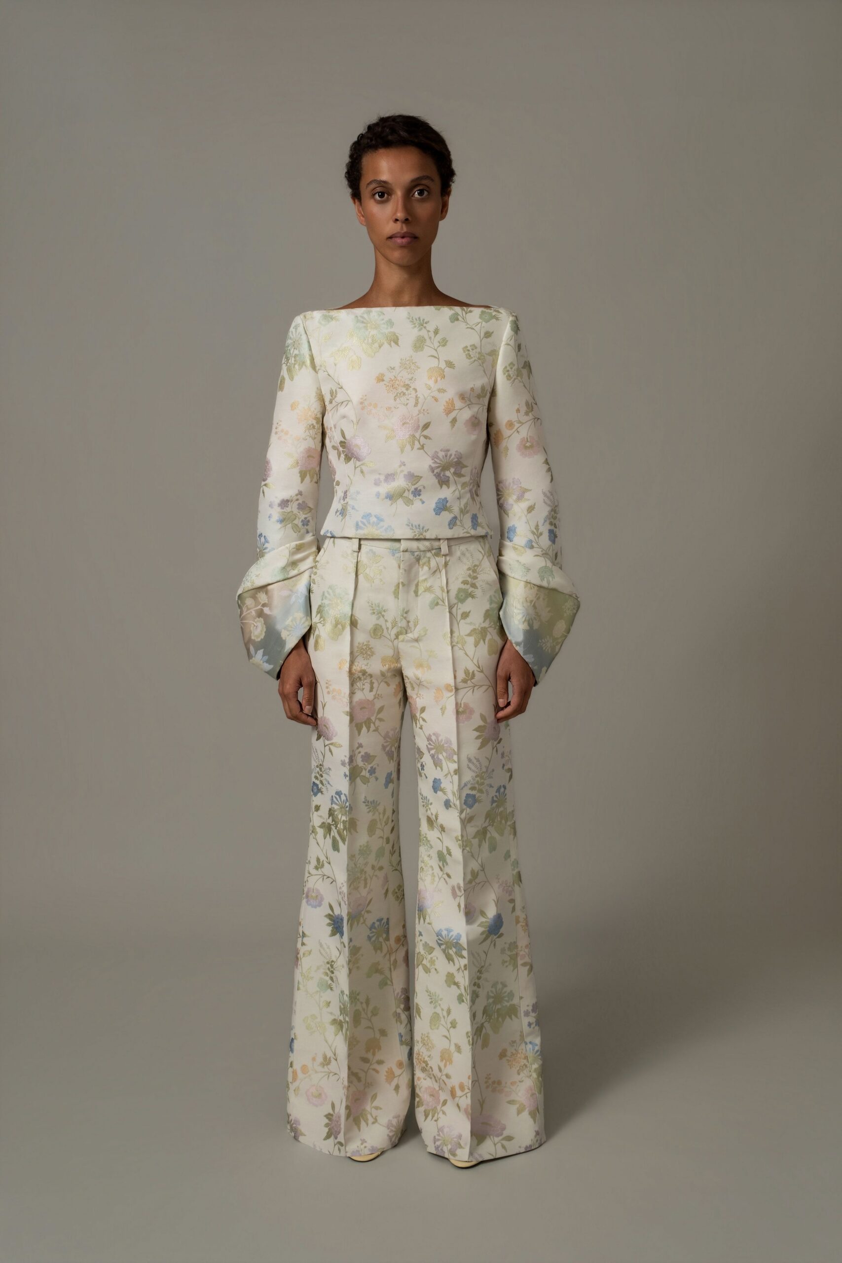 Cream satin top printed with washed wildflowers, sleeves opening into turned-back trumpet cuffs. Matching high-waisted flares in the same floral satin, pressed creases sharp down the front. Pale pointed toes just visible under the hem. Registry office at noon, then cake in a garden gone slightly overgrown.