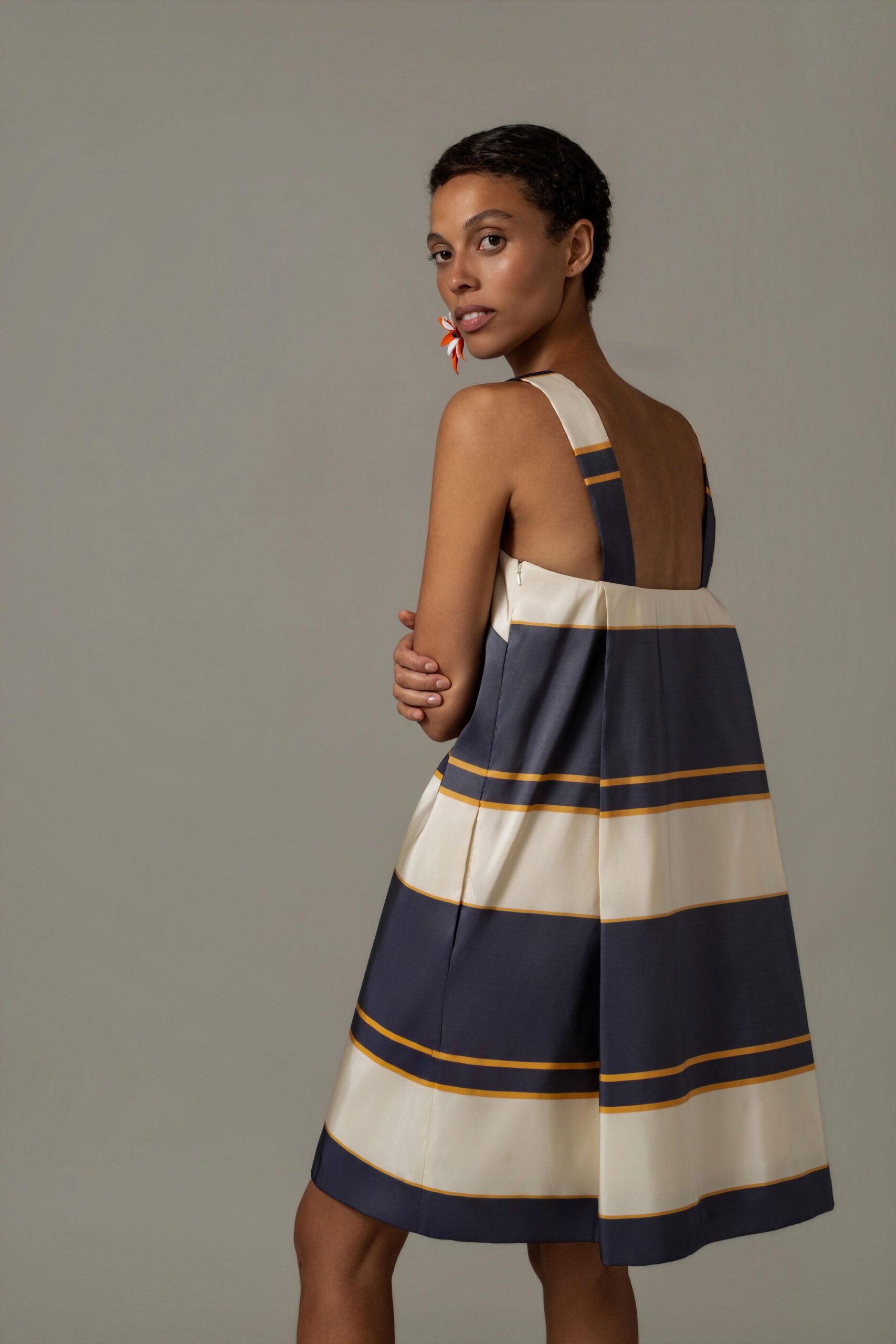Orange and white petal earring, oversized against the jaw. Cream and slate striped sundress in crisp cotton, box pleat falling from the back zip. Afternoon veranda light, somebody has just brought out iced tea.