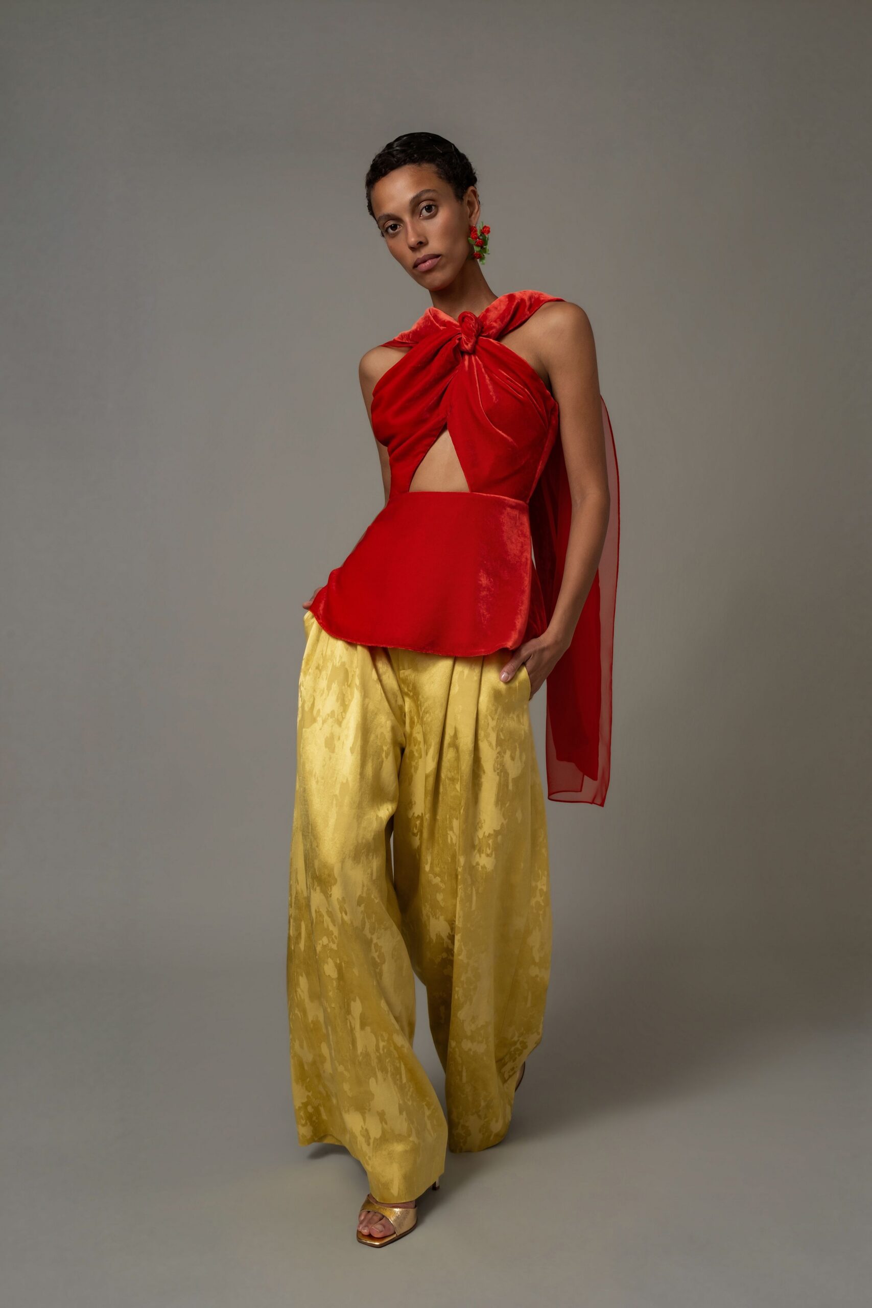 Scarlet satin halter top knotted at the throat, with a sheer back drape. Wide ochre jacquard trousers, washed with cloudy tonal patches. Gold sandals with a squared toe. Walking into a wedding lunch after the church bells have stopped.