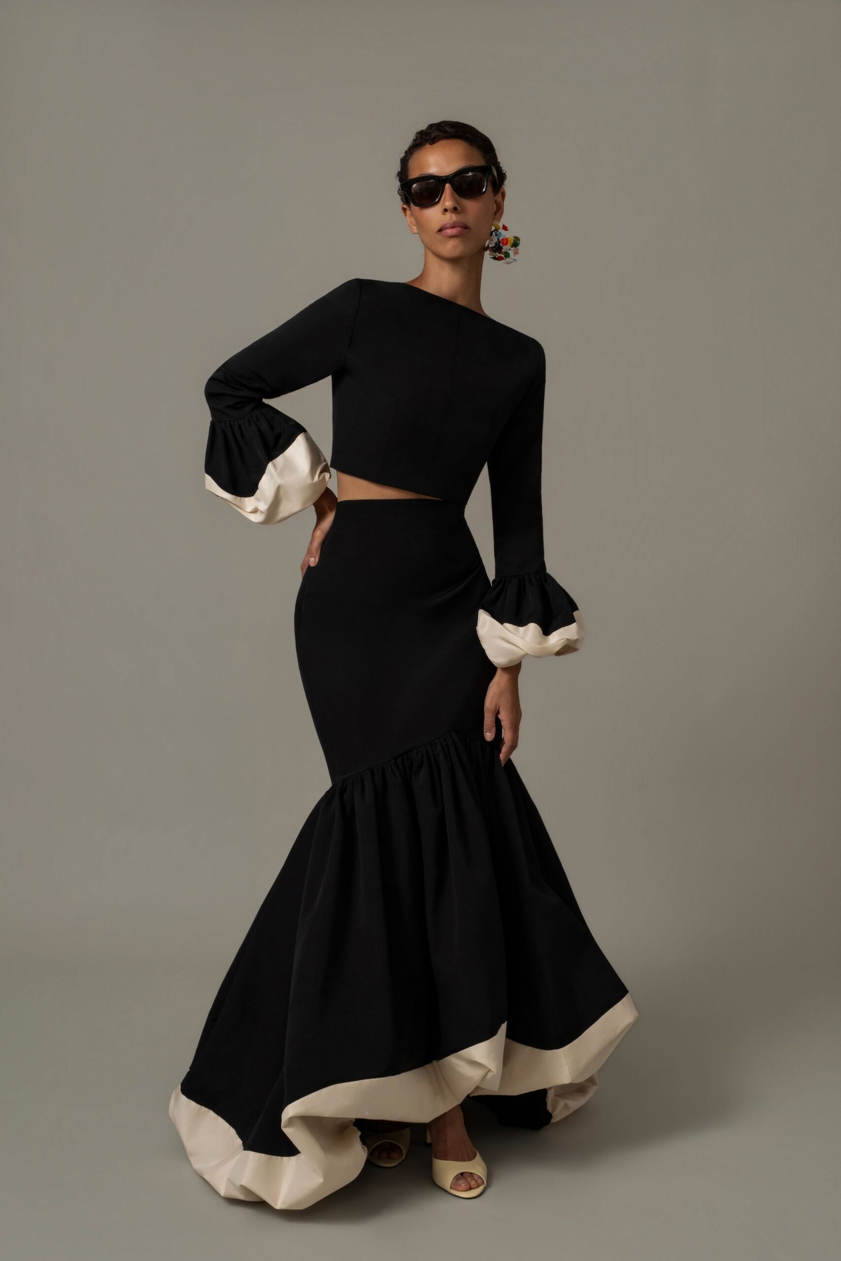 Black cat-eye sunglasses with thick glossy frames. Black cropped top with a bateau neck and puffed ivory cuffs. High-waisted black mermaid skirt, ruffled low and faced in ivory. Ivory peep-toe mules. For arriving late to a courtyard wedding after the vows are done.