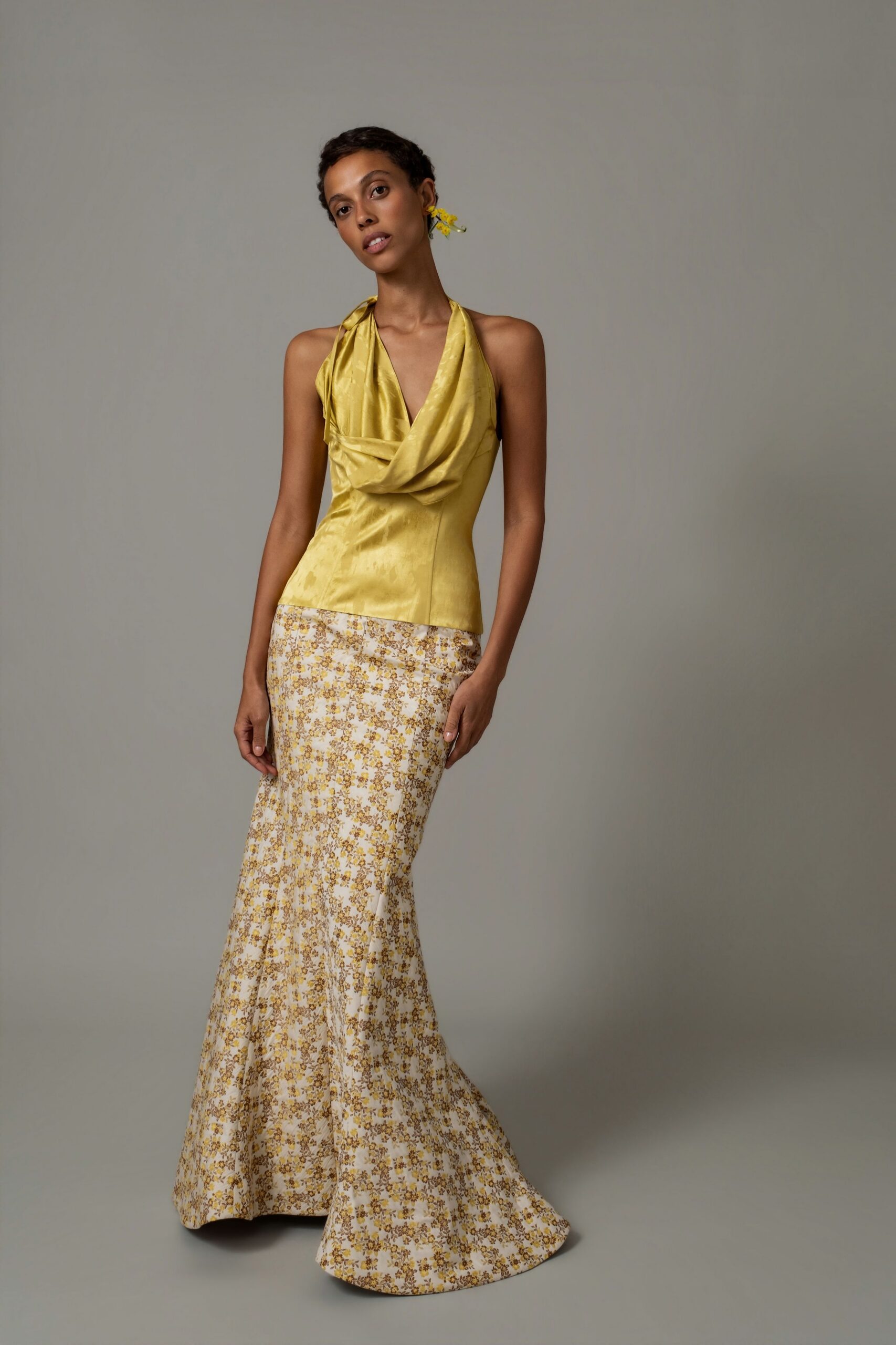 Chartreuse satin halter top with a soft cowl folding low at the chest. Cream bias-cut maxi skirt scattered with tiny ochre and brown flowers. Yellow blossom cluster at one ear. Garden wedding before the chairs are set straight.