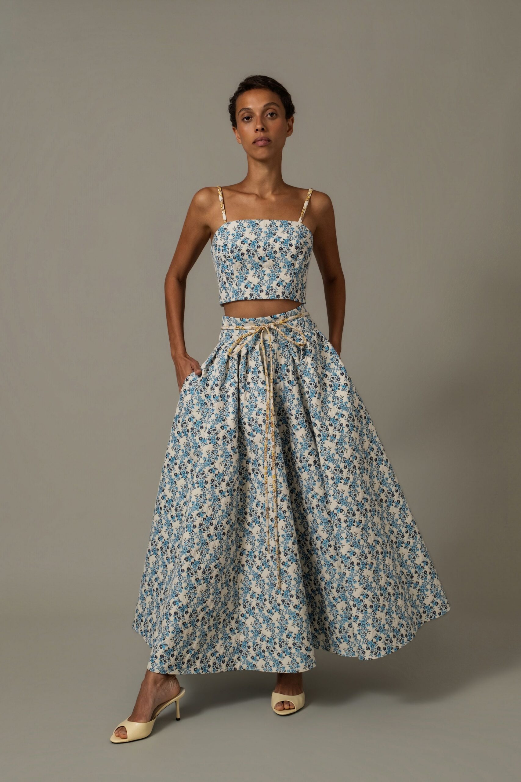 White cotton crop top scattered with small blue sprig florals. Full matching skirt, gathered at the waist with a trailing cord tie. Buttercream open-toe mules on slim stiletto heels. Walking through a garden wedding before the chairs have been set out.