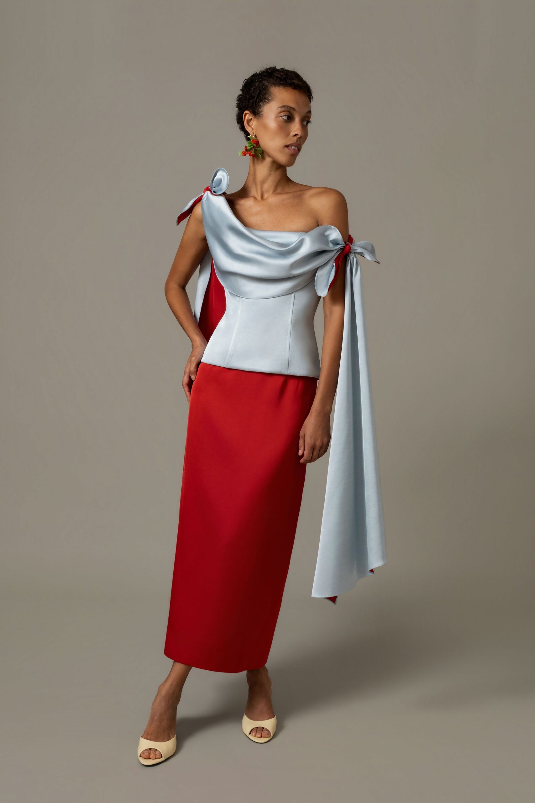 Cherry-cluster earrings with glossy green leaves. Ice-blue satin top, cowl neckline tied into shoulder bows with trailing sash. Column skirt in tomato red crepe, cut clean and close. Buttercream leather mules with a squared open toe. Garden wedding reception, someone has just carried out the cake.