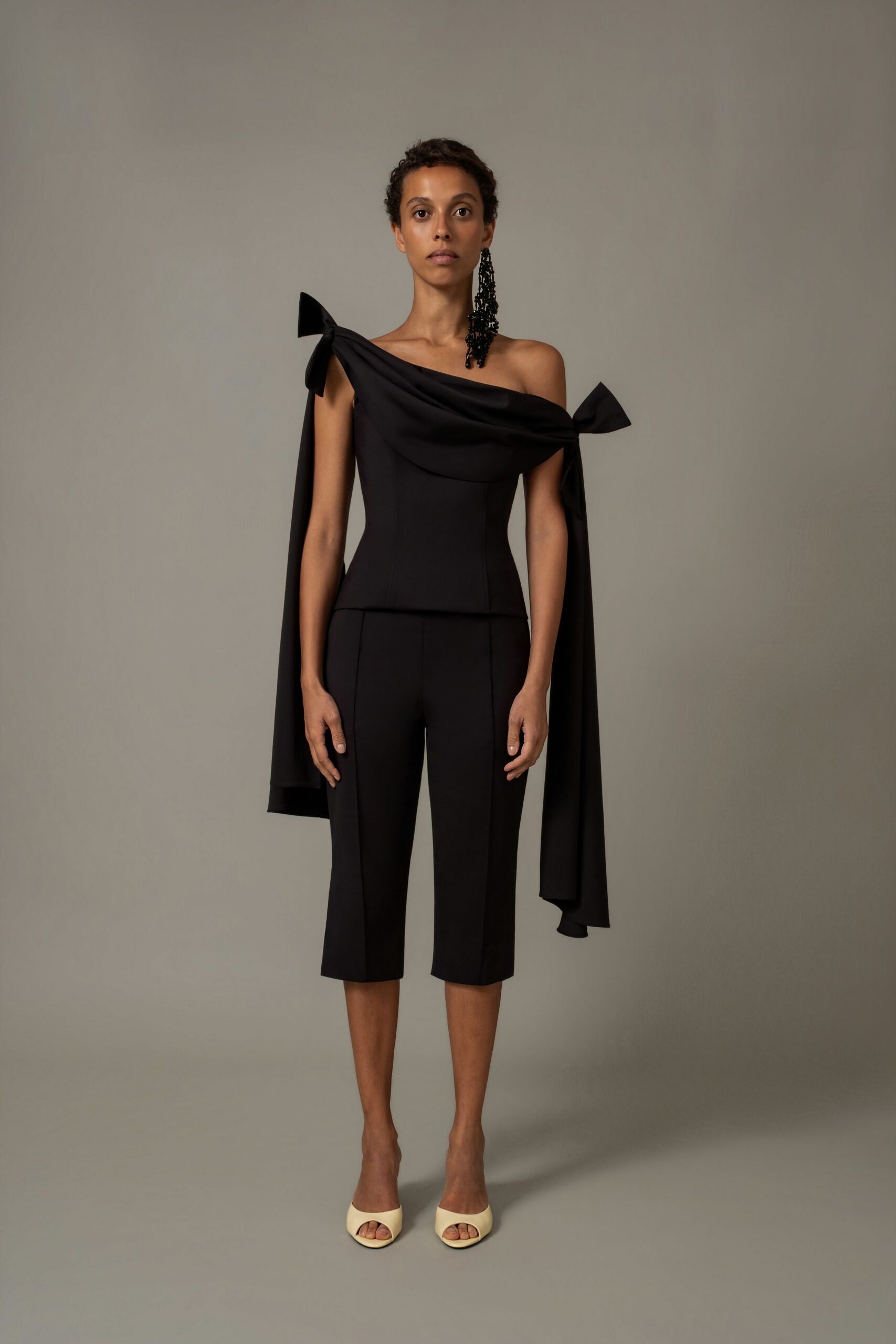 Black crepe top with a folded off-shoulder band and long bow streamers. Matching black cropped trousers, pressed seams running straight to mid-calf. Black beaded tassel earring brushing one shoulder. Cream leather peep-toe mules. For slipping into a recital hall after the doors have closed.