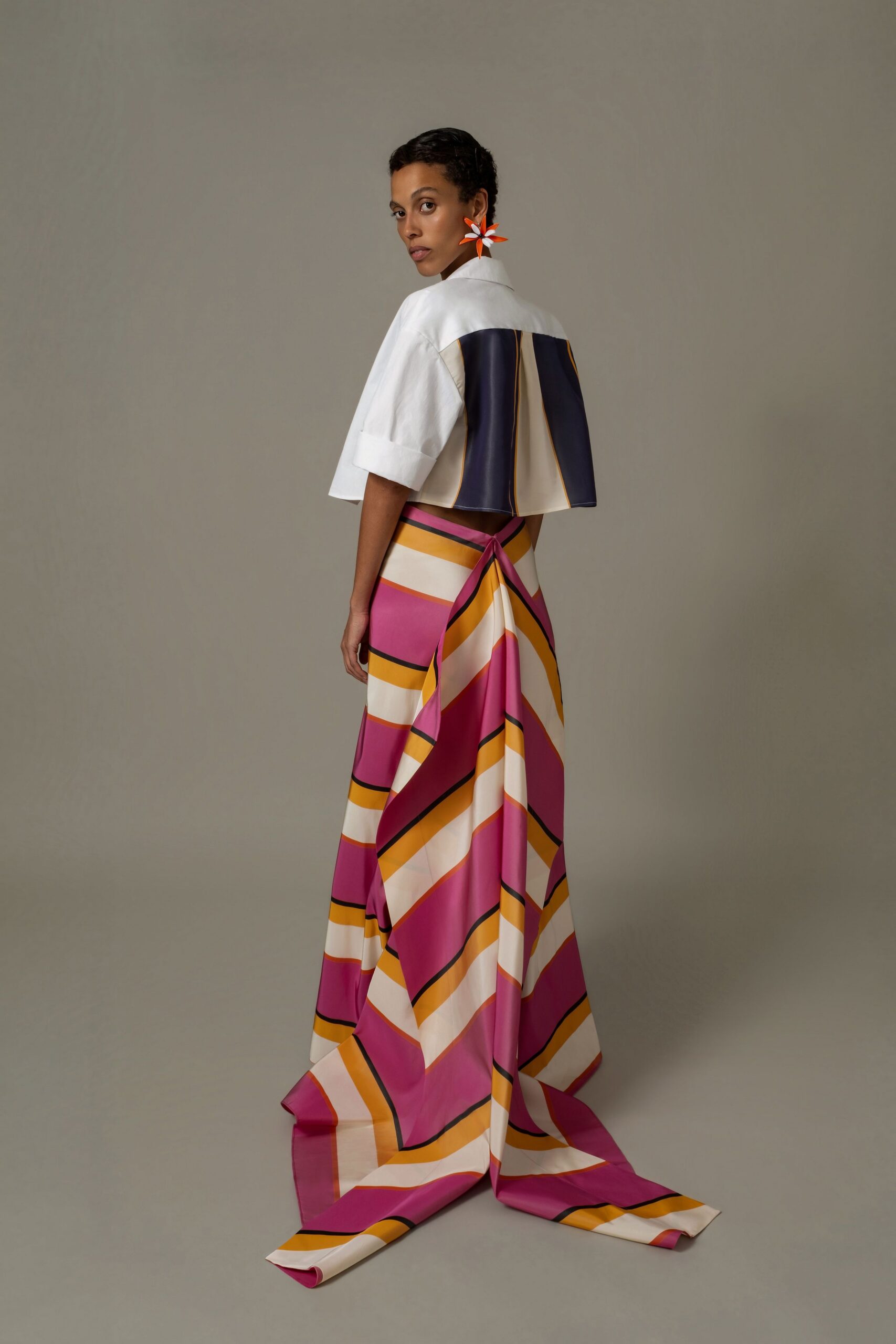 Oversized orange flower earring with white-tipped petals. White structured crop jacket, back paneled in navy and cream. Bias-cut skirt in magenta, cream and marigold stripes, knot draped at the back. Walking out to a terrace lunch above the harbor, tablecloths snapping in wind.