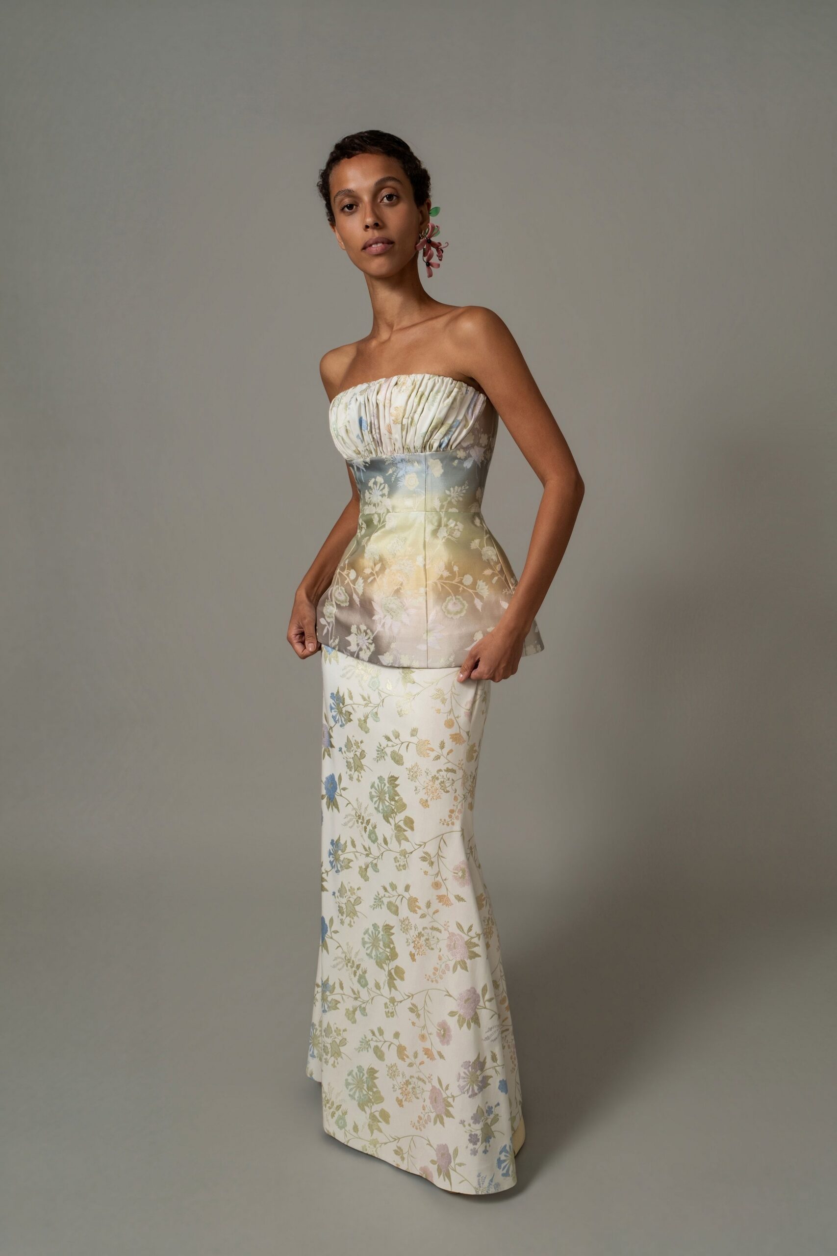 Pink petal cluster earring with two small green leaves. Strapless floral satin bustier, ruched ivory panel above a flared peplum. Column skirt in matching satin, scattered with faded blue and straw blossoms. Garden wedding, the hour when the chairs are being folded away.