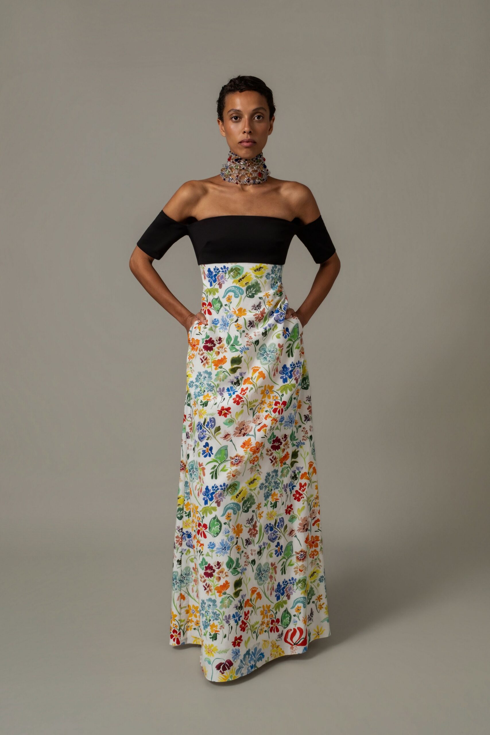 Tall collar of clear faceted beads dotted with colored stones. Black bandeau bodice with off-shoulder sleeves over a white floral satin column, pockets opening at the hip. Garden wedding lunch gone on past the last bottle of white.