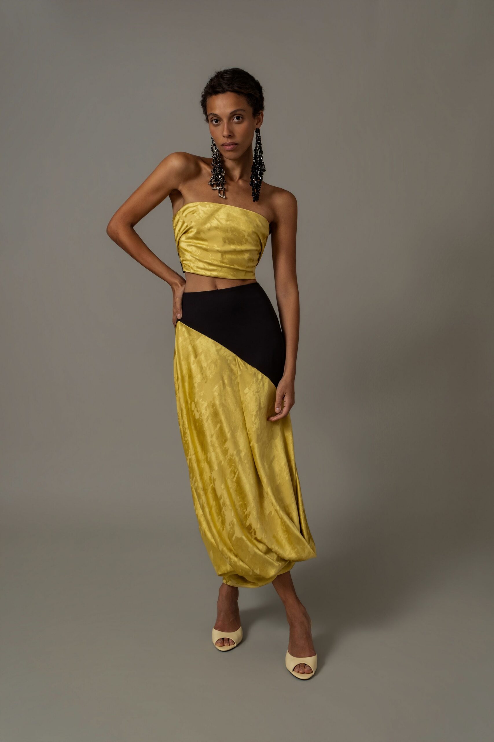 Long black beaded tassel earrings brushing the collarbone. Strapless citron jacquard bandeau, softly wrinkled across the bust. Bias-cut skirt in matching jacquard, black yoke slicing down the hip. Butter peep-toe mules. For crossing a hotel atrium after midnight with your shoes still on.