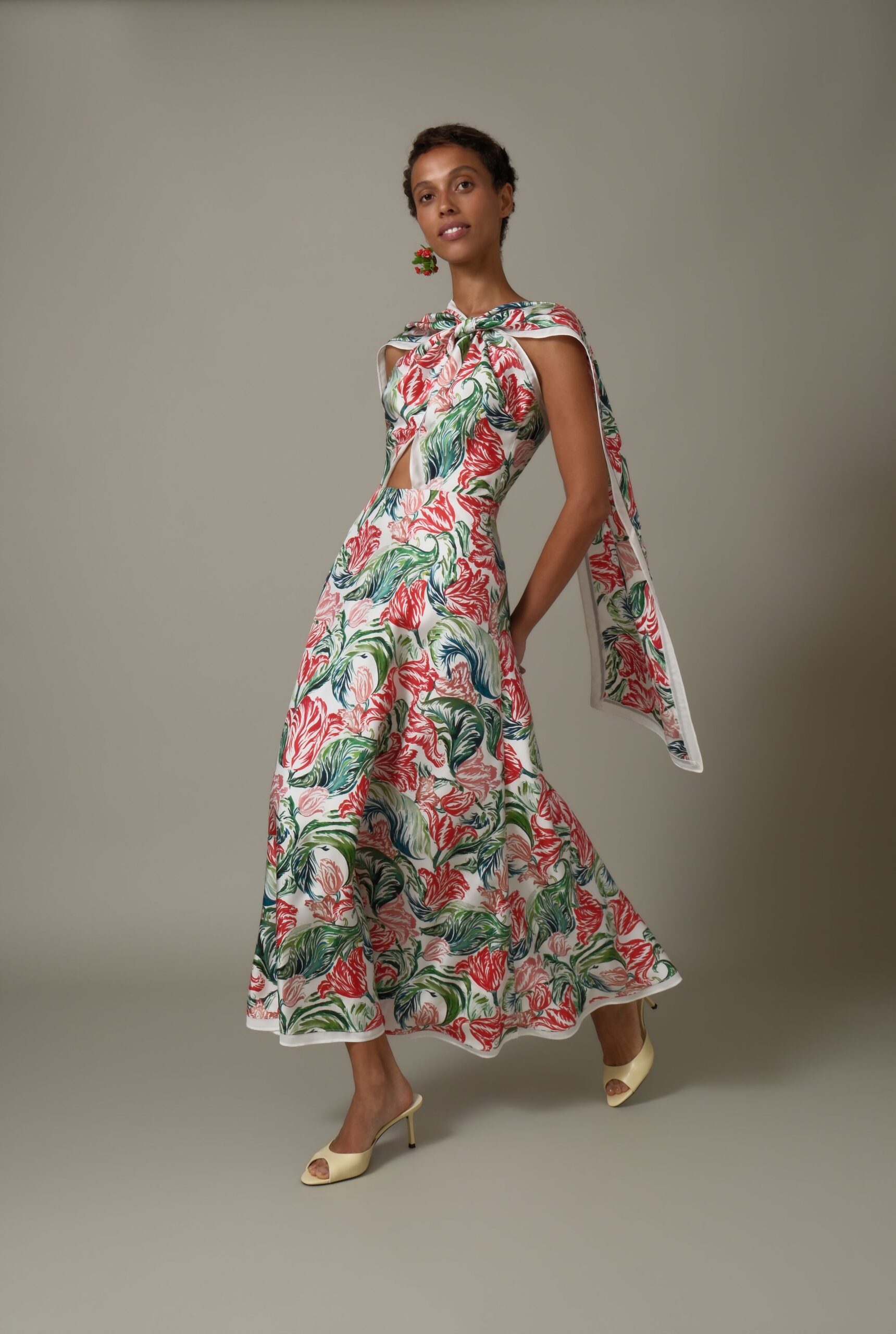 Cherry-cluster drop earring in glossy red and green beads. White cotton dress printed with painterly tulips, neck twisted into a capelet drape. Buttercream satin mule sandals with a slim stiletto heel. Garden wedding lunch, someone has just moved the tables into shade.