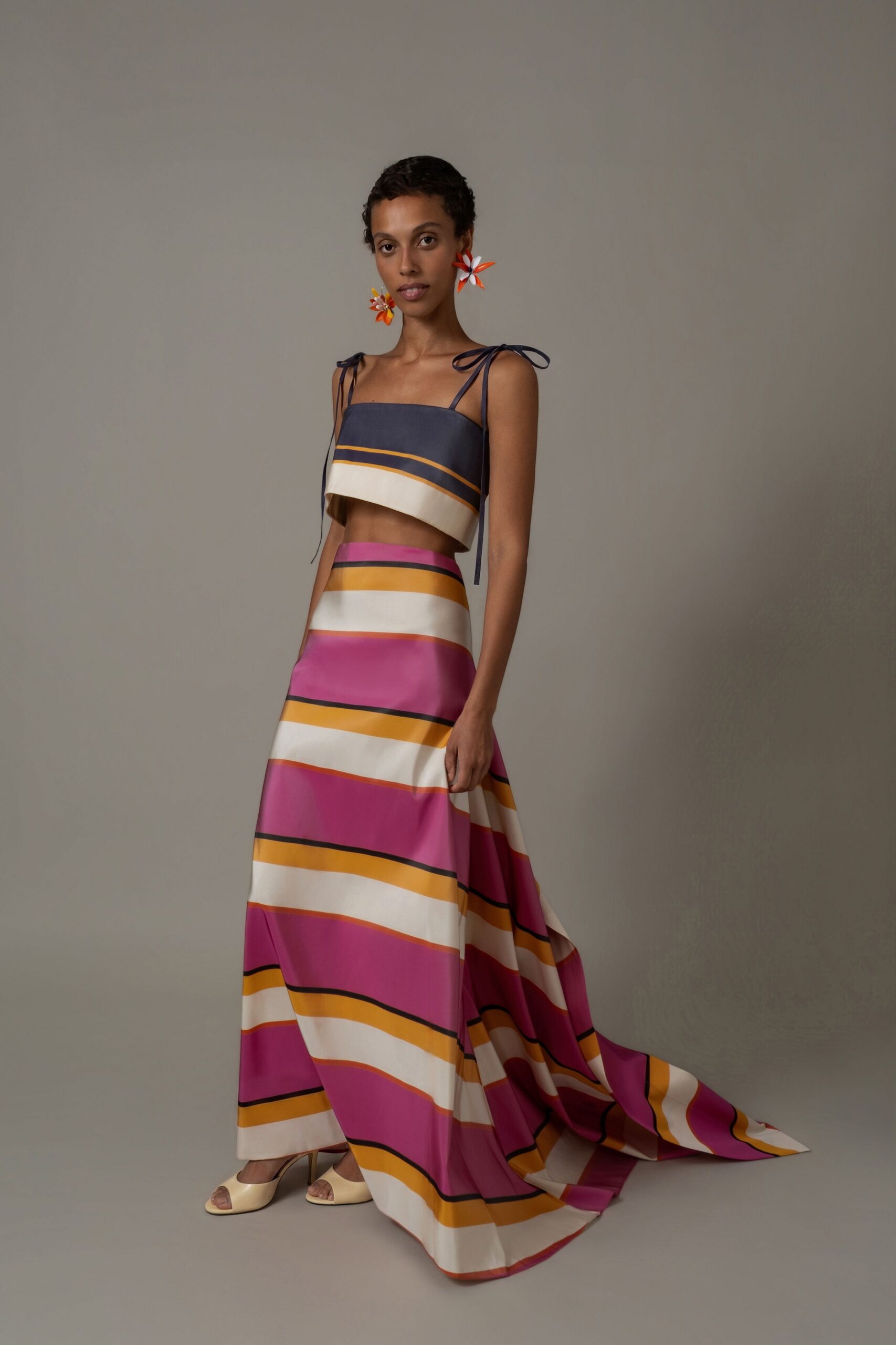 Oversized pinwheel flowers in orange, white and yellow enamel. Slate satin crop top with shoulder ties and a dipped front hem. Bias-cut satin skirt in magenta, cream and marigold deck stripes. Butter peep-toe mules with a low curved heel. Walking the hotel veranda before breakfast, sea air lifting the train.