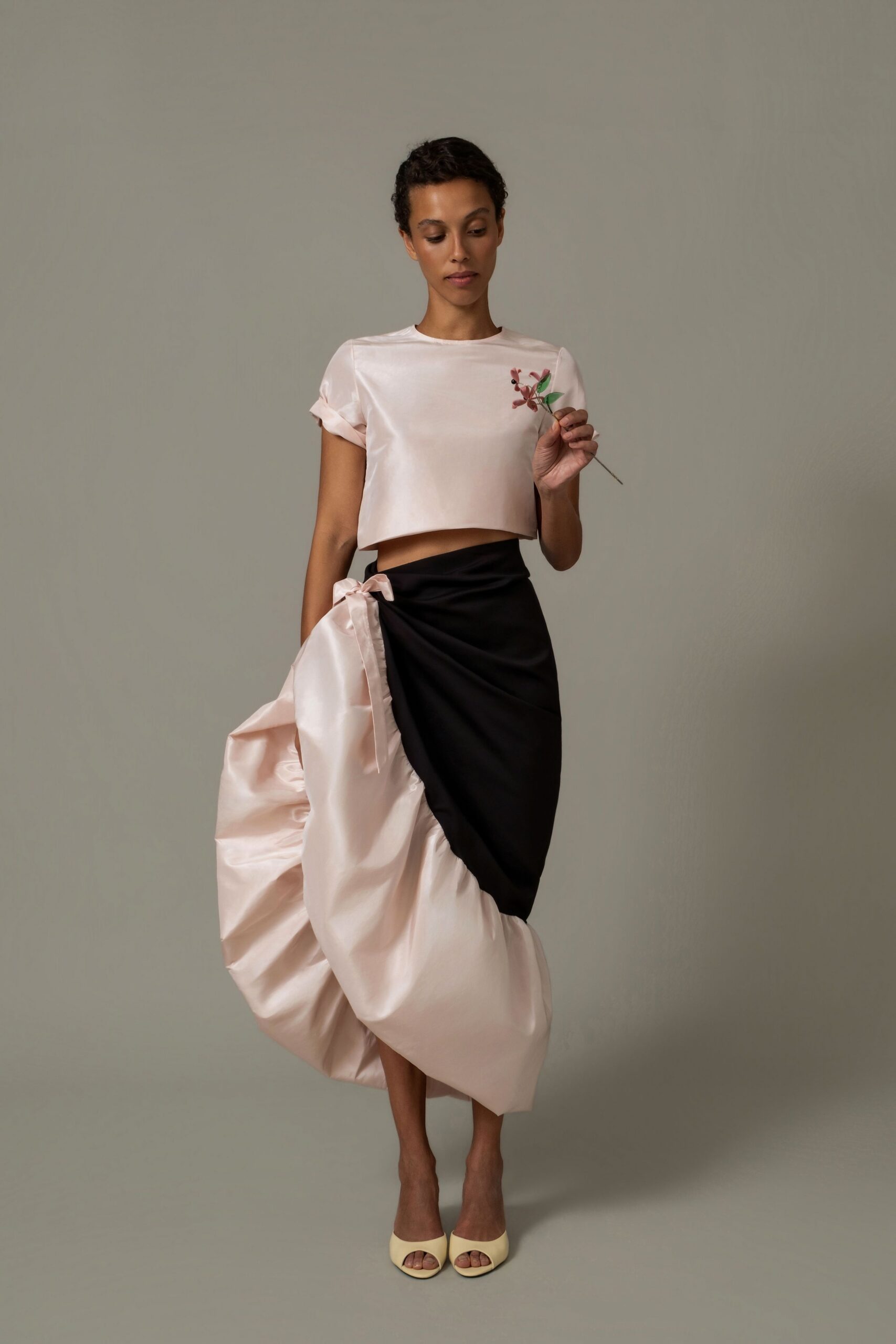 Blush satin cropped top with turned cuffs and a clean bateau neck. Black wrap skirt over a pale shell satin bubble underlayer, tied at the hip. Buttercream peep-toe mules. Holding one stem from the centrepiece after everyone else has left.