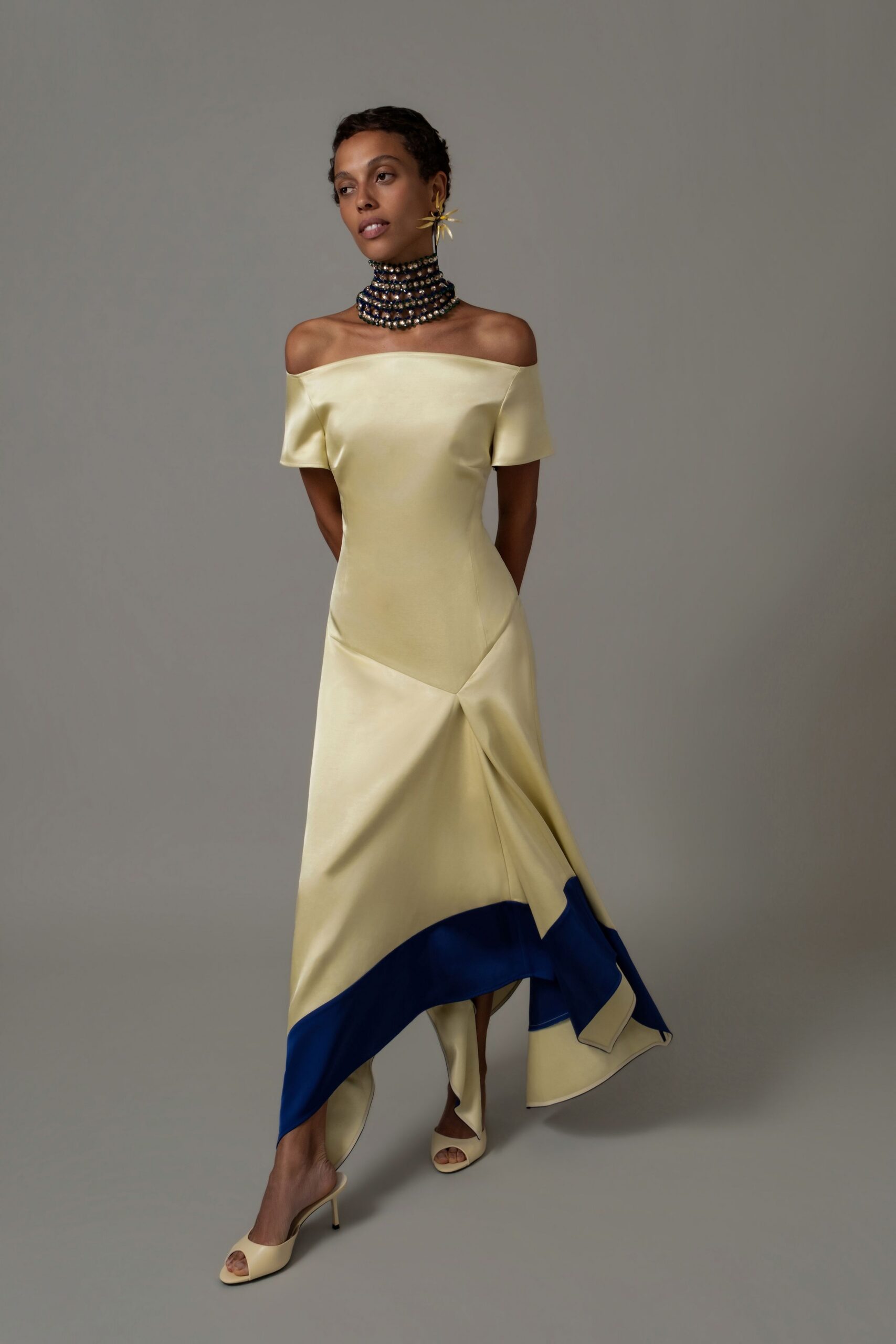 Stacked navy and pearl chokers rising high under the jaw. Spiked gold starburst earring at one ear. Butter satin off-the-shoulder gown, twisted at the hip into an uneven cobalt-lined hem. Butter satin mule sandals with slim stiletto heels. Crossing a museum atrium after the string quartet has packed up.