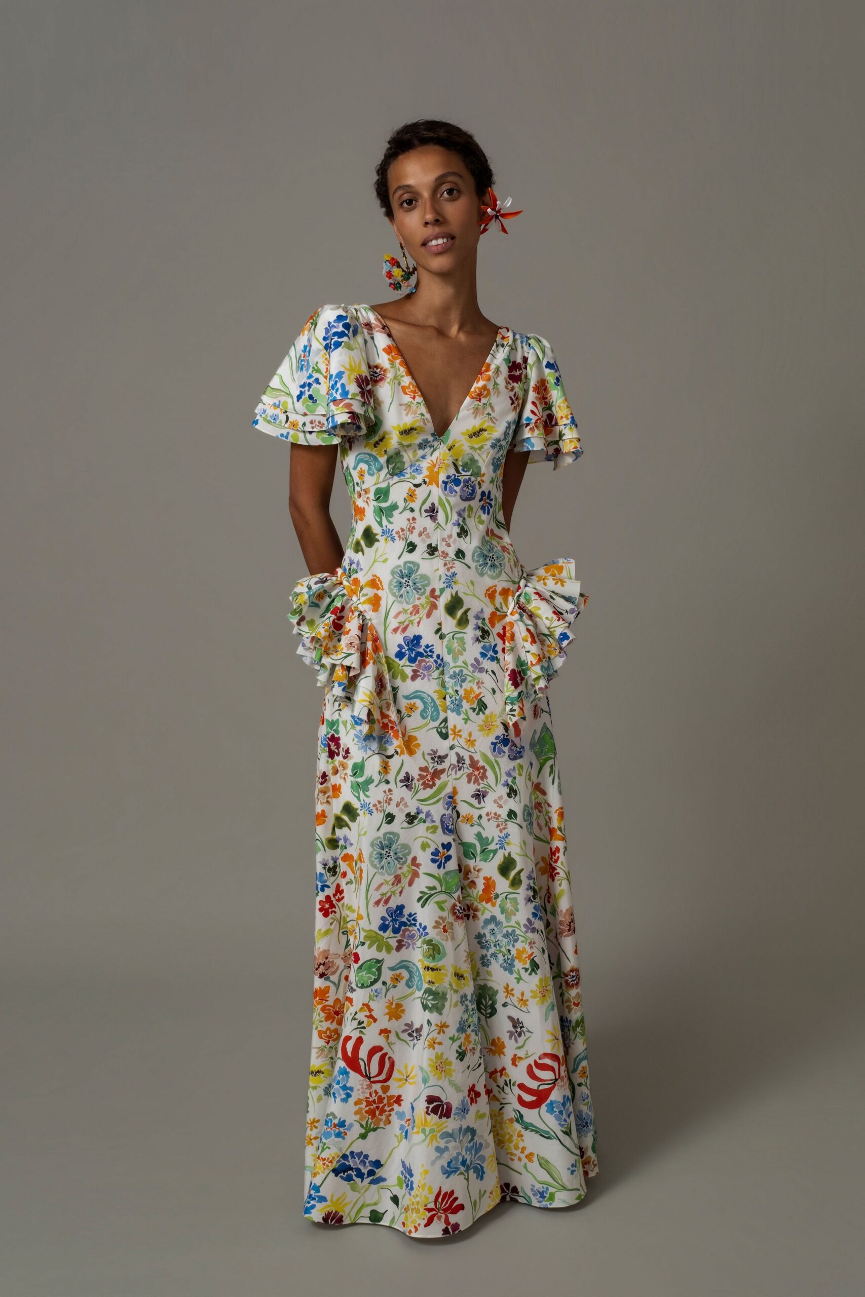 Mismatched floral drop earrings, one clustered and one pinwheel-bright. White floral cotton gown with flutter sleeves and tiered hip ruffles. Garden wedding reception, someone has moved the tables onto the grass.