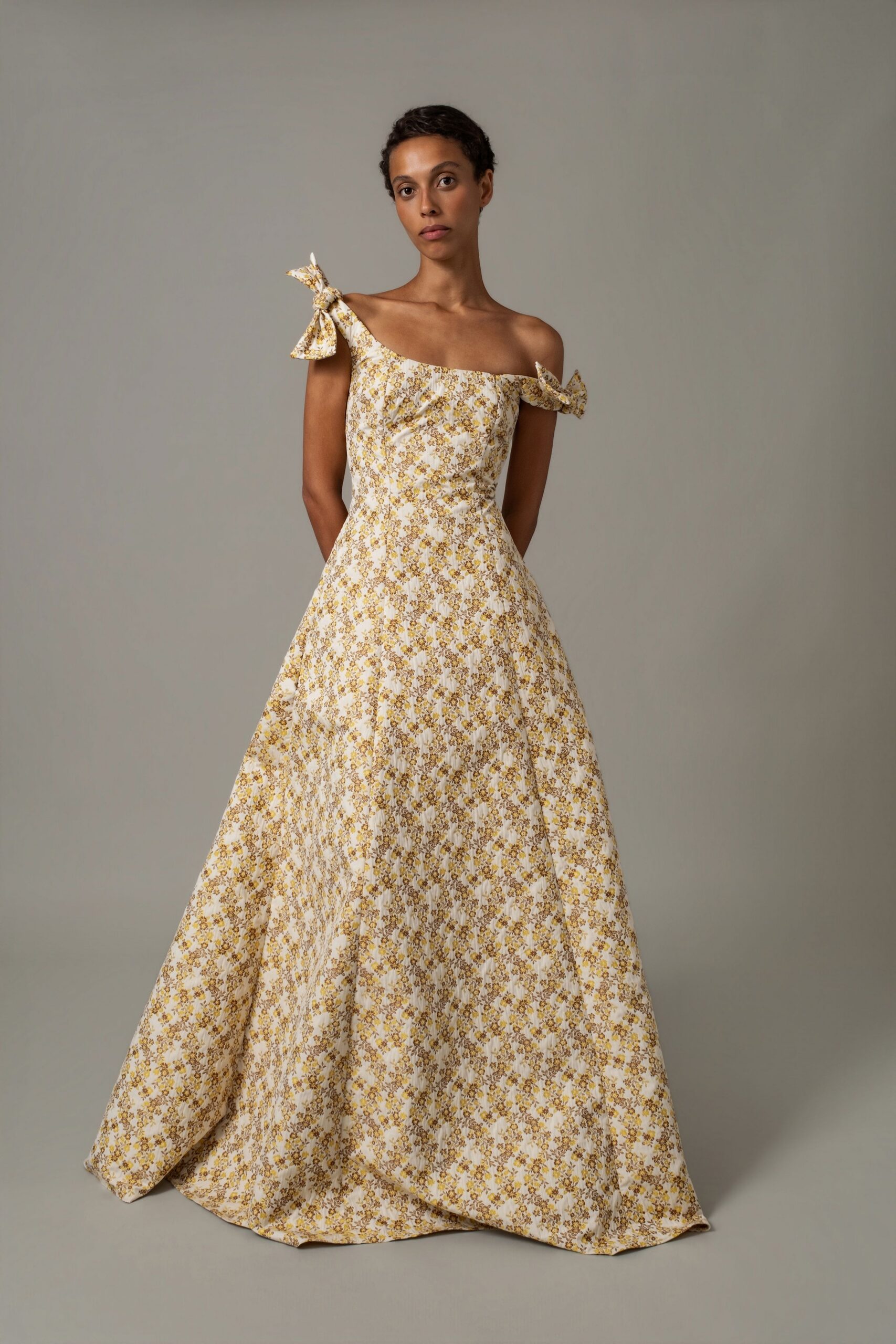 Off-shoulder ivory gown in yellow sprig cotton, bow-knotted straps and paneled skirt. Garden wedding after the chairs have sunk slightly into the lawn.