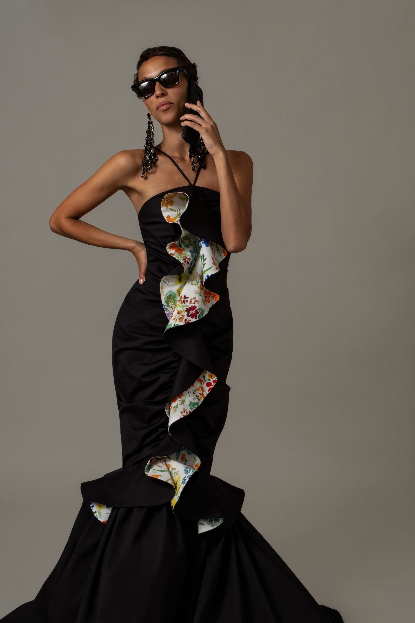 Black cat-eye sunglasses with thick glossy rims. Black beaded chandelier earrings brushing the shoulders. Strapless black satin gown, floral lining flashing through a deep serpentine ruffle. Fine black cord crossing the collarbones. Taking a call in the museum lobby before the benefit doors open.