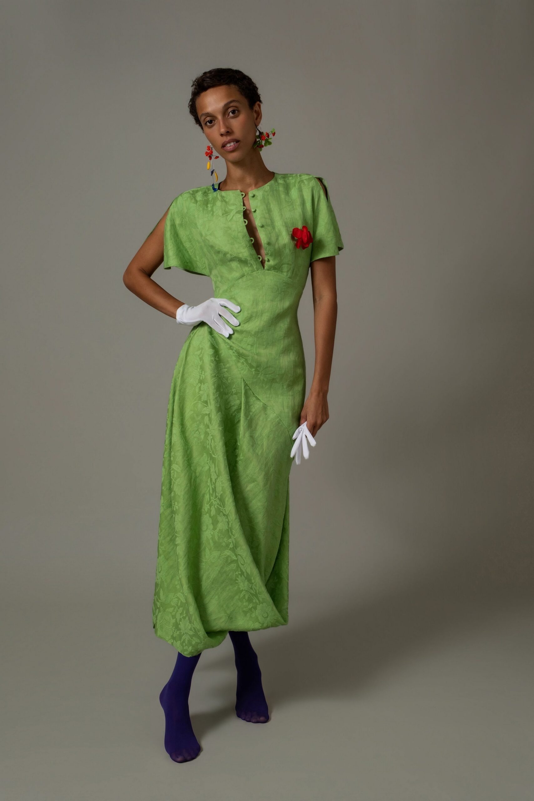 Apple-green jacquard dress with cape sleeves and looped buttons opening at the chest. Red flower pinned high on the bodice. White wrist gloves, one carried loose by the fingertips. Opaque violet tights. For a garden wedding where the grass is still wet from rain.