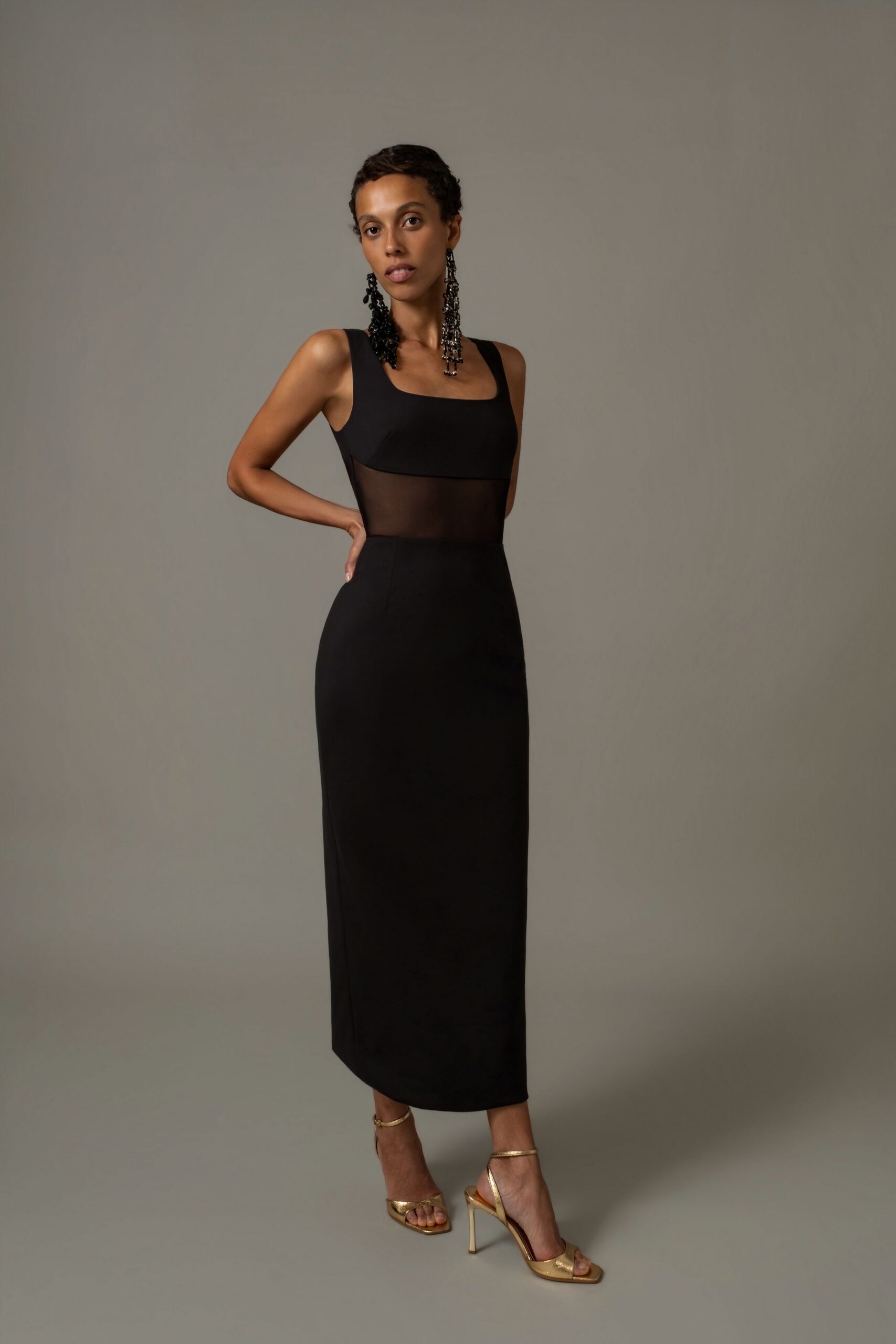 Long black beaded fringe earrings grazing the collarbone. Black column dress with a square neckline and sheer mesh waist panel. Gold ankle-strap sandals with slim stiletto heels. For a hotel bar wedding where the candles are already burning low.