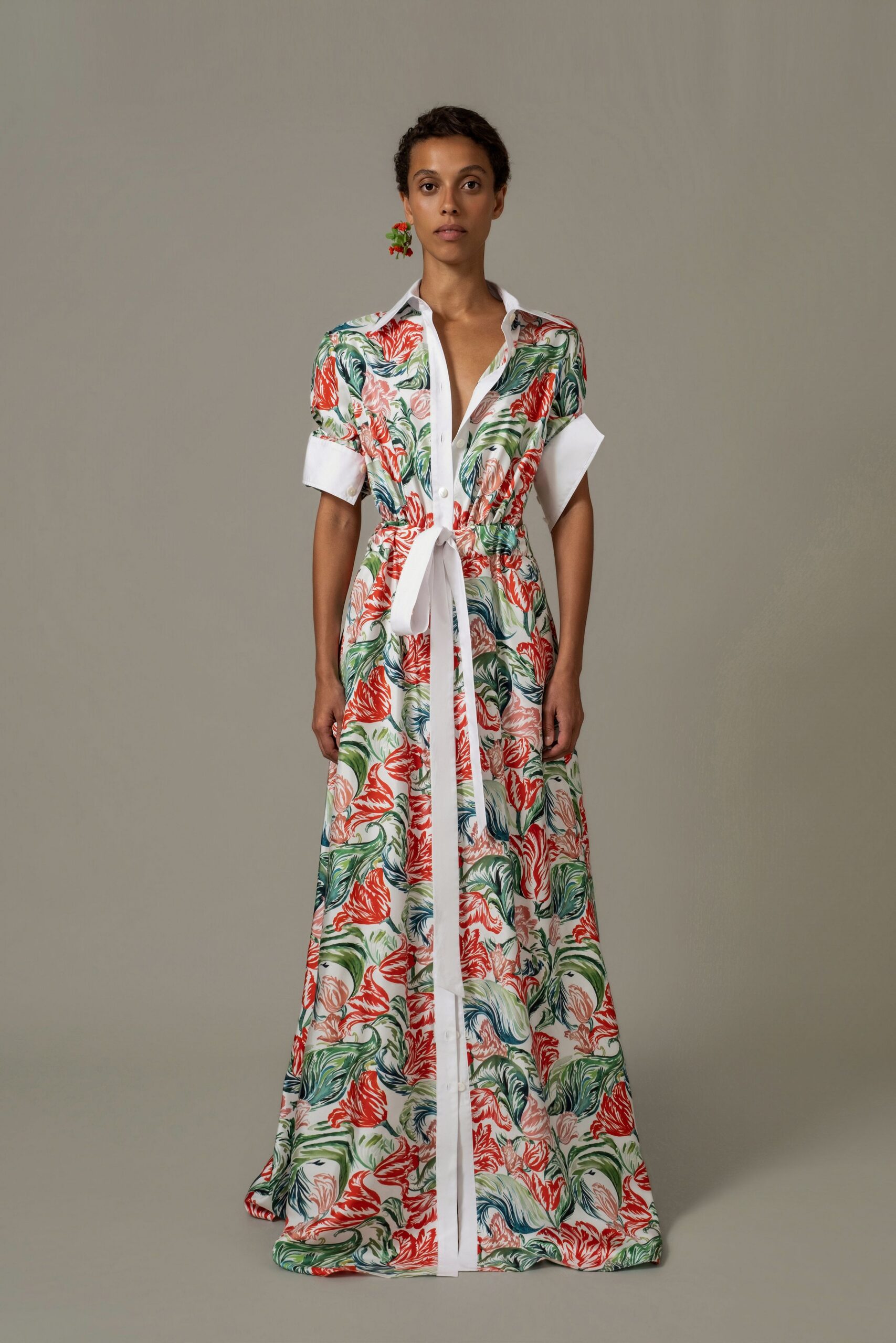 Single drop earring with clustered red blossoms. White shirtfront maxi in painterly coral-and-leaf print, cuffs rolled broad. Long self-tie sash knotted low, one tail falling to the hem. Garden wedding breakfast, already warm enough to leave two buttons undone.