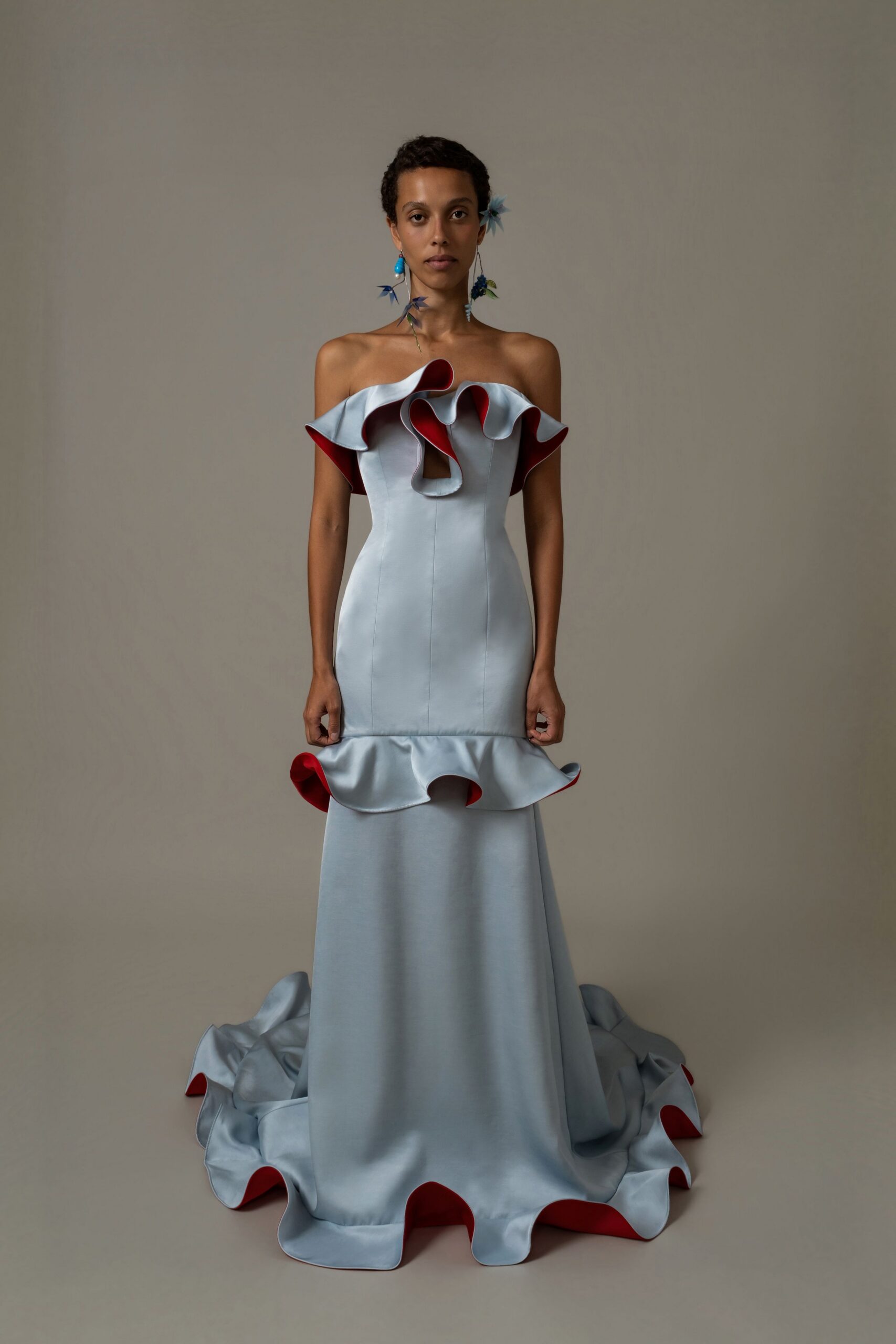 Blue floral drop earrings with long bead-tipped stems. Powder-blue satin column gown, scarlet-backed ruffles circling neckline, hip and train. Garden wedding after the heat breaks, champagne already warm on the table.