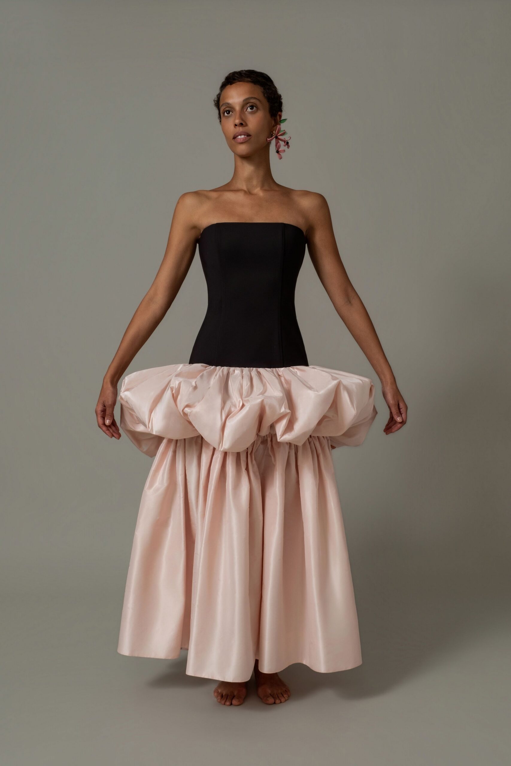 Strapless black bodice dropping into a blush taffeta bubble overskirt. Pink lily sprig earring trailing behind one ear. Standing barefoot on the drawing-room rug after everyone else has gone.
