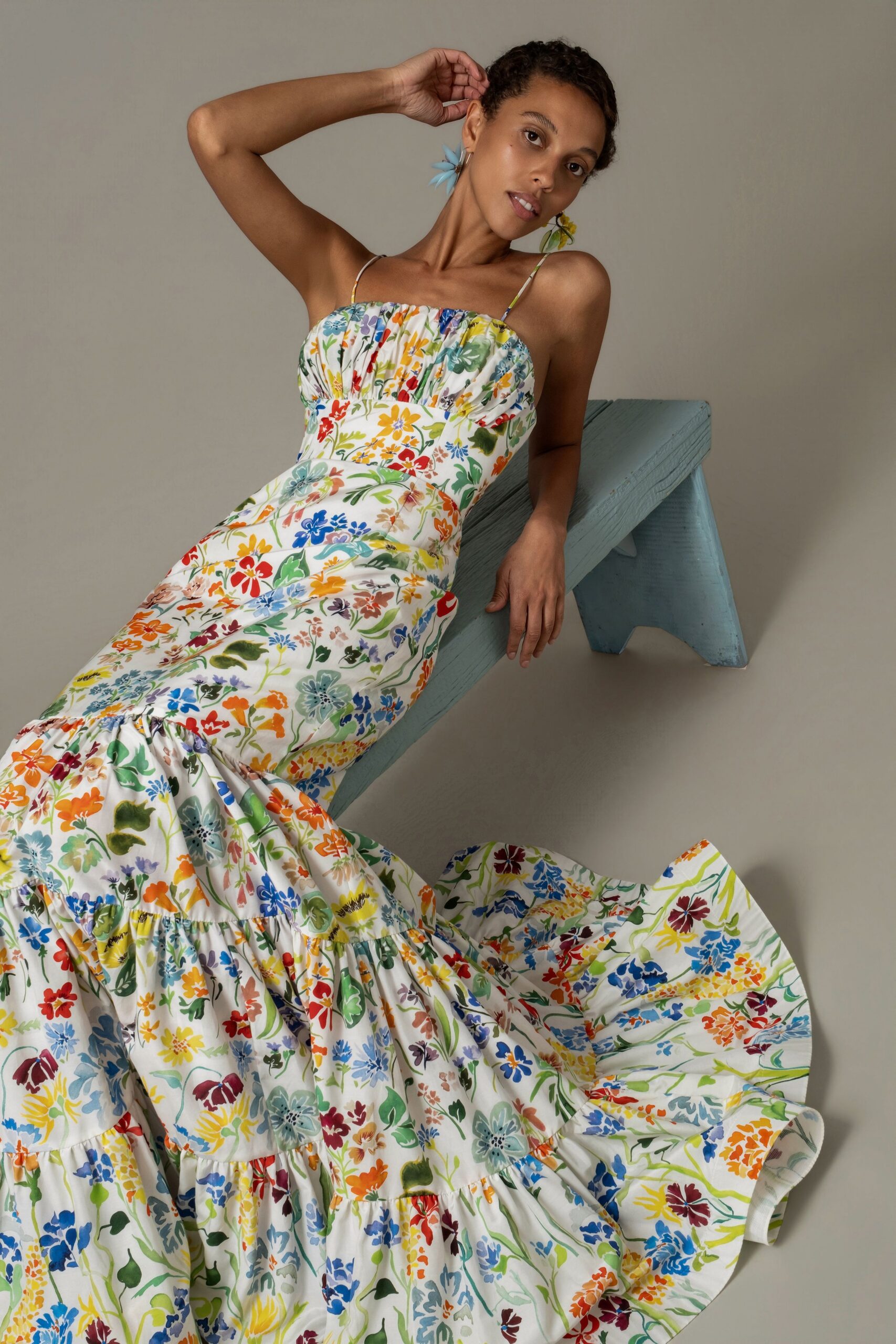 White cotton sundress scattered with wildflower print, ruched bodice and tiered hem. Mismatched flower-drop earrings, one pale blue and one yellow. Garden wedding spillover, sitting on the porch after the cake is gone.