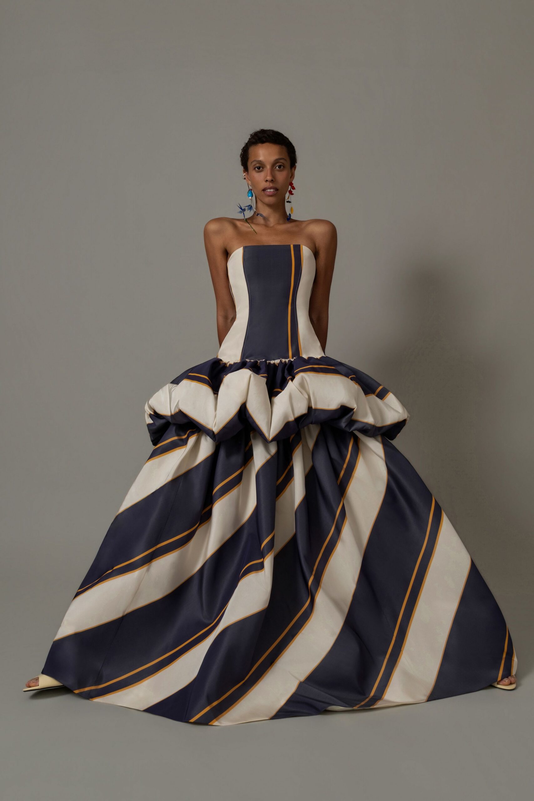 Strapless ballgown in crisp navy-and-ivory faille, peplum puffed into pointed folds. Mismatched flower drops brushing the collarbone. Pale open-toe sandals peeking from the hem. At the museum gala after midnight, standing under the whale skeleton.