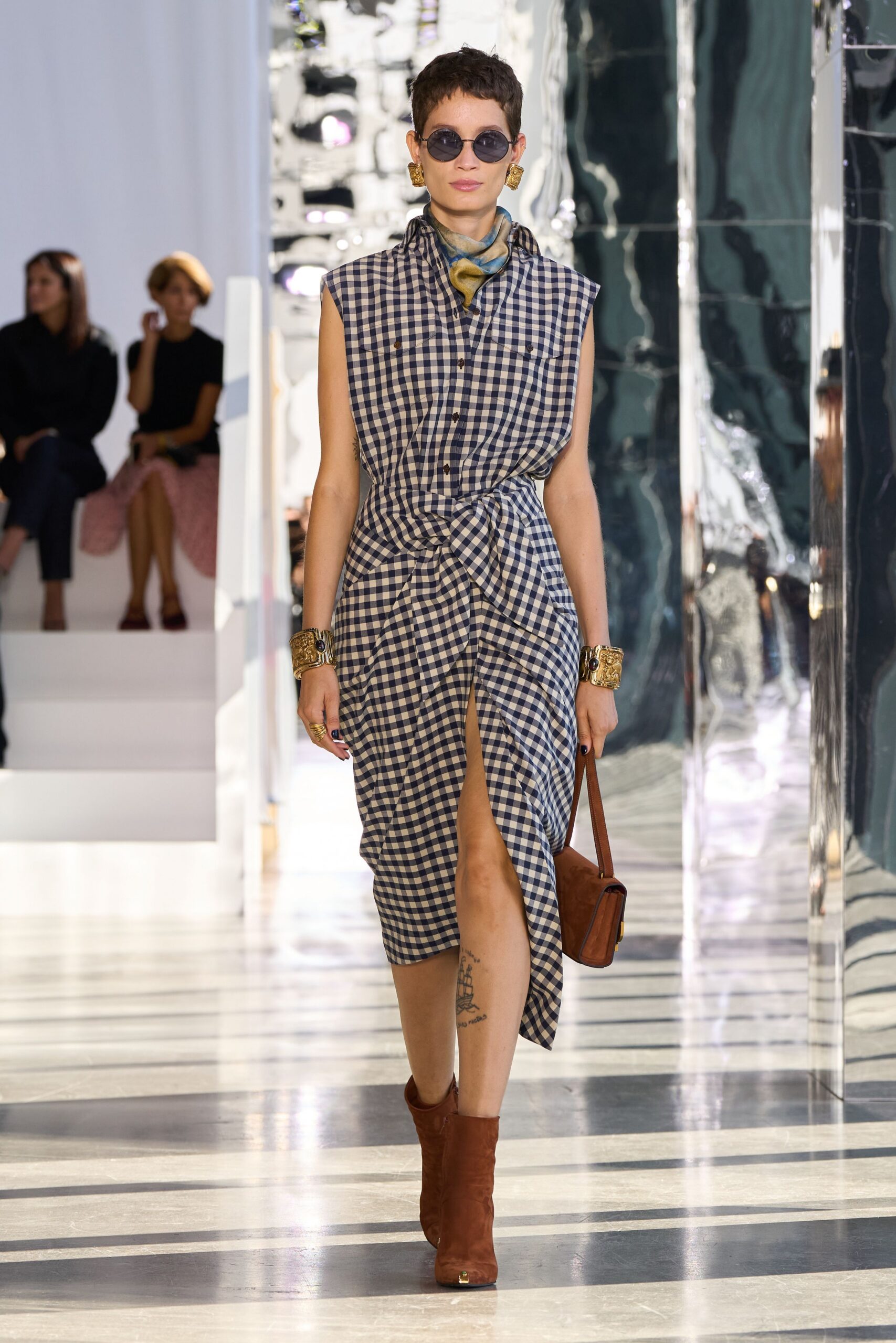 Round smoke lenses in thin dark rims. Blue and ochre silk twisted close at the throat. Navy gingham shirtdress, cut sleeveless and knotted hard across the waist. Cognac shoulder bag in brushed suede. Slouchy chestnut suede boots with a tiny gold toe cap. Walking into a seaside hotel lobby before lunch service starts.