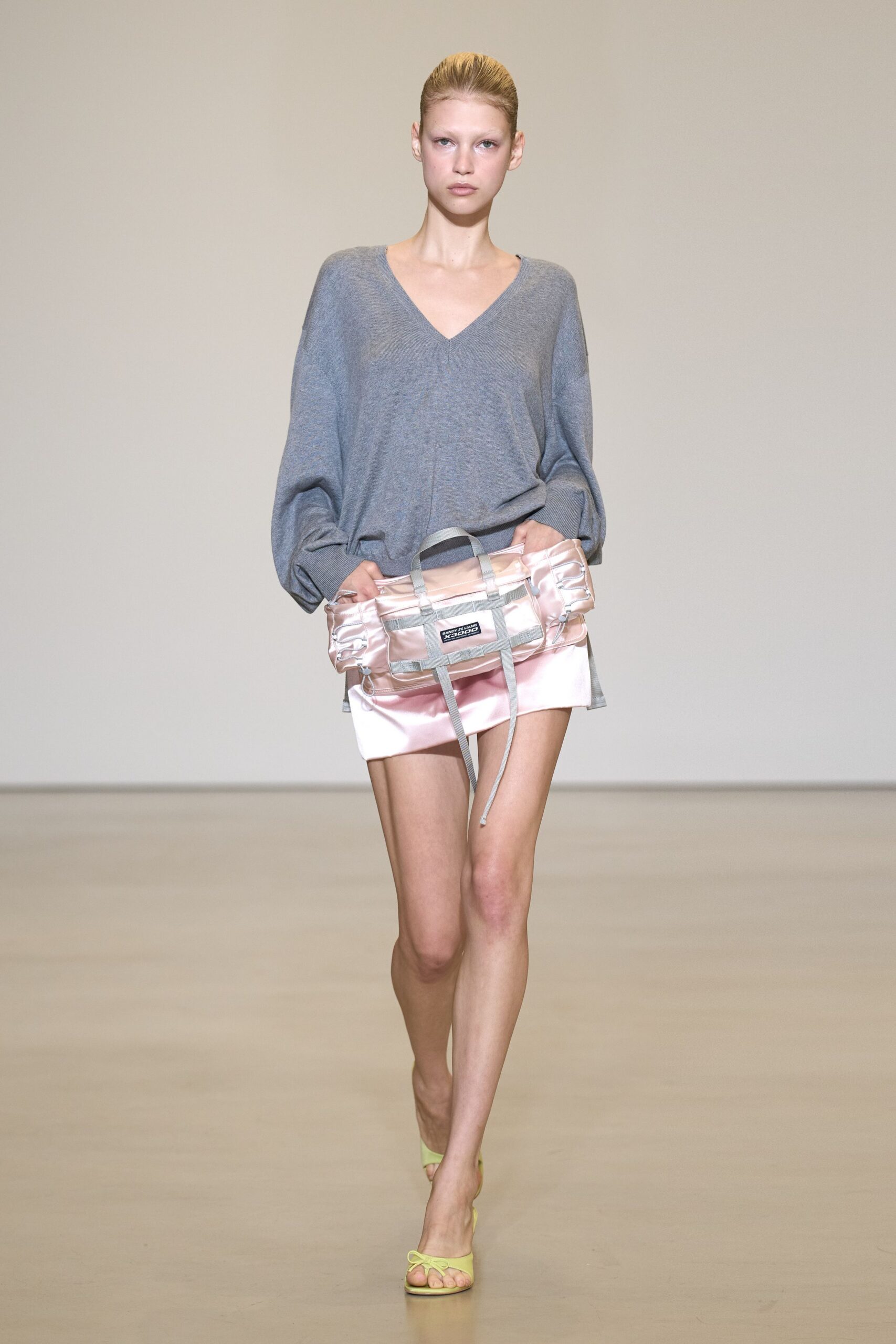 Slouchy heather-grey knit sweater with a deep V and dropped shoulders. Blush satin duffel with webbing straps and a loose cord end. Pale pink satin mini, smooth and slightly crumpled at the hip. Chartreuse thong mules with thin lace-up ties. For buying flowers at closing time, still carrying your gym bag.