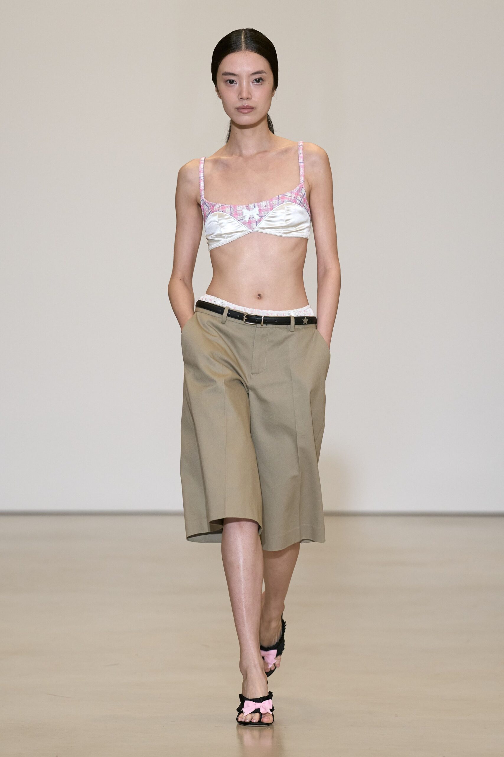 Ivory satin cups under a pink plaid yoke, tiny bow centered. Stone cotton twill culottes cut below the knee with pressed creases. Narrow black belt with a small silver star by the buckle. Black fuzzy sandals topped with pale pink bows. Walking back from a corner shop in borrowed clothes and yesterday's eyeliner.