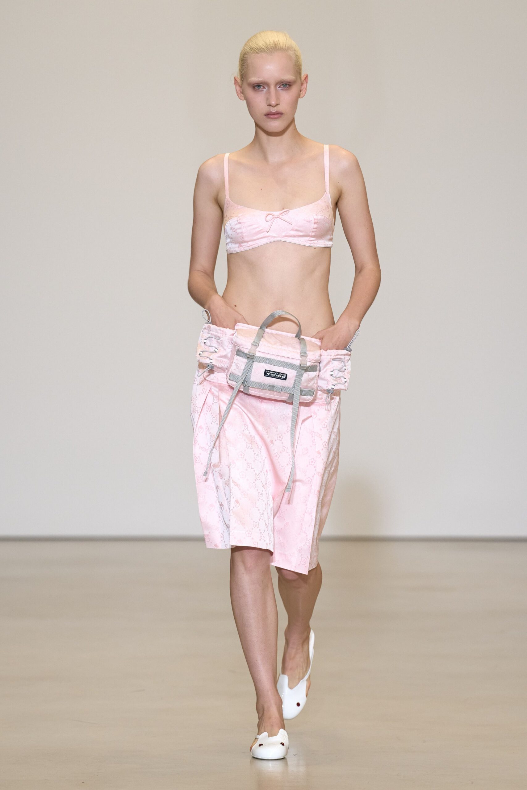 Pale pink satin bralette with a tiny bow at center front. Low-slung pink satin wrap skirt, monogram faintly washed across it. Utility belt bag in matching satin, webbing straps dangling past the hem. White backless mules with blunt rounded toes. For buying cold medicine at the corner shop still dressed from bed.
