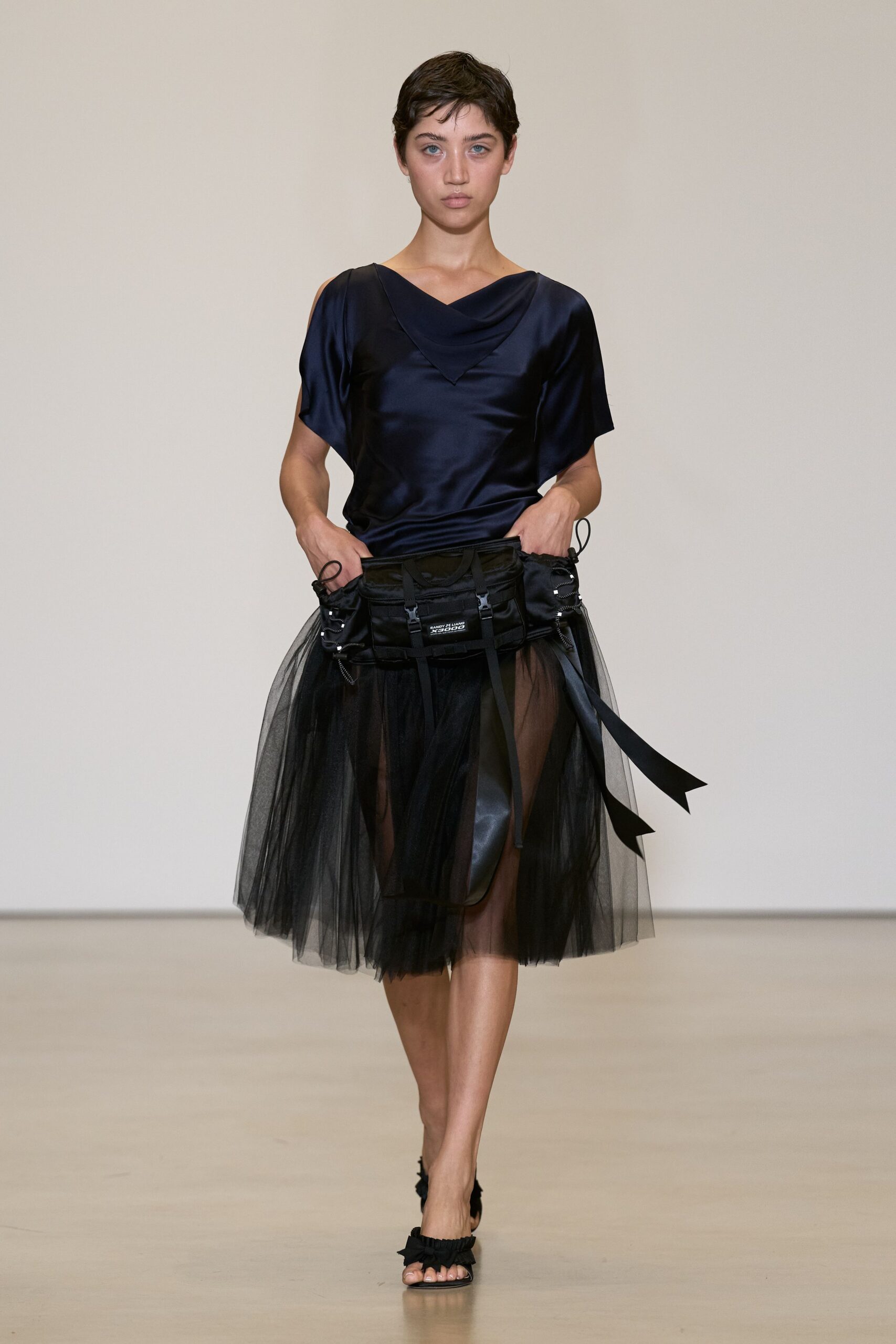 Ink satin top with a draped cowl folding to a point. Black tulle skirt, sheer layers floating over a shorter underpanel. Nylon cargo belt bag with side pouches and dangling webbing straps. Flat sandals with ruffled black frills at the toes. Walking the long corridor to a hotel ballroom after midnight teardown.