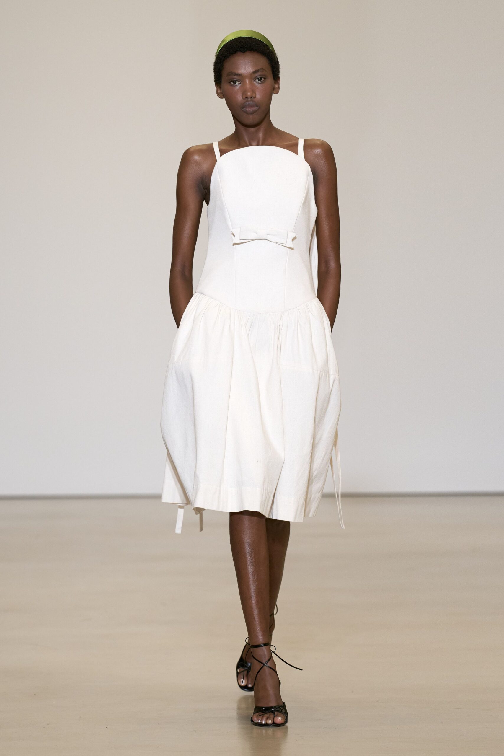 Pistachio satin headband, smooth against cropped hair. Ivory cotton dress with a curved bib front and tiny waist bow. Black lace-up sandals, cords winding thinly around the ankle. Garden wedding morning, before the chairs have been unfolded.