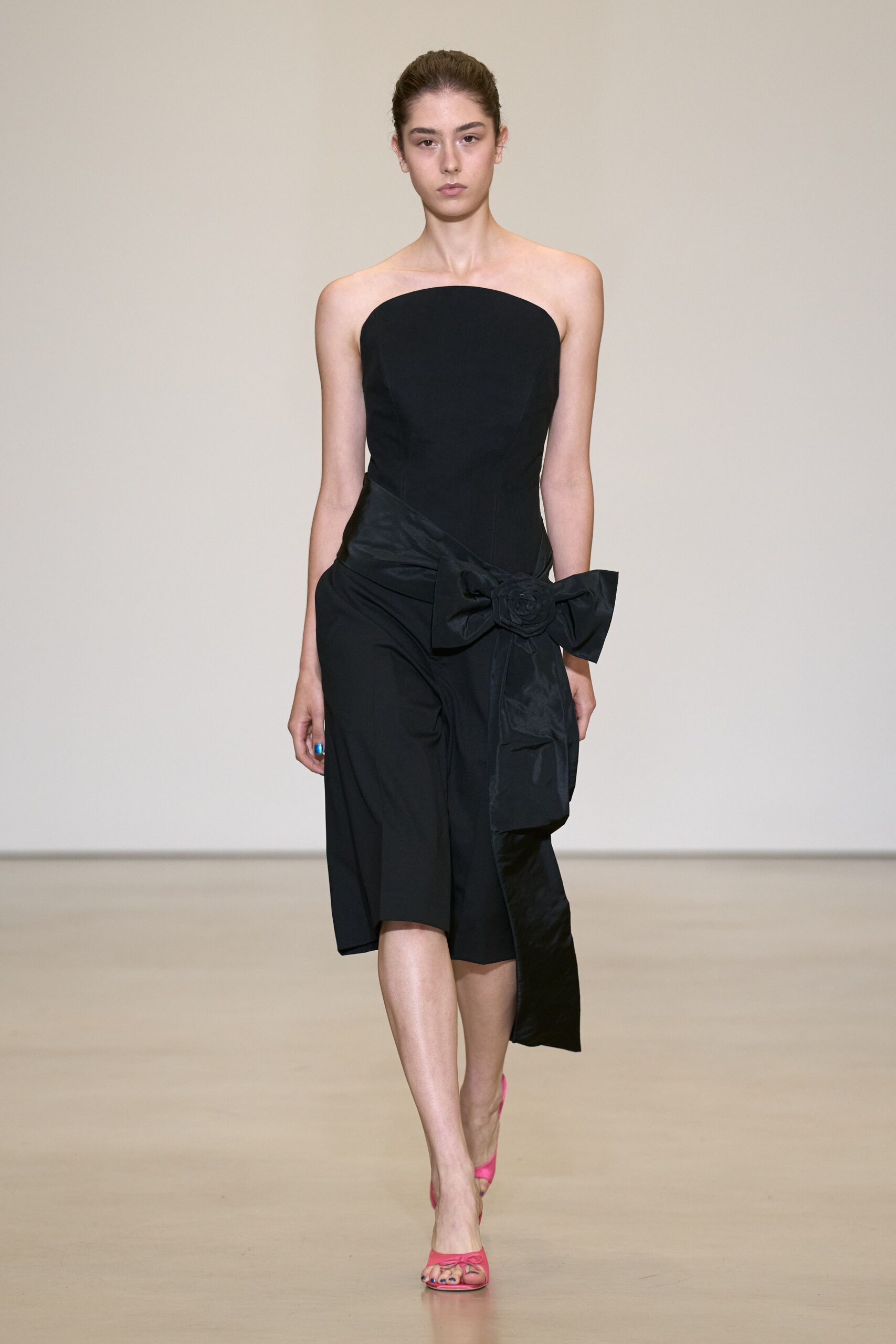 Black strapless dress with a curved neckline and sculpted princess seams. Crisp taffeta sash knotted at the hip, rosette centered in the bow. Hot-pink satin mules with a small bow over the toes. Hotel ballroom after midnight, carrying your heels by the second dance.