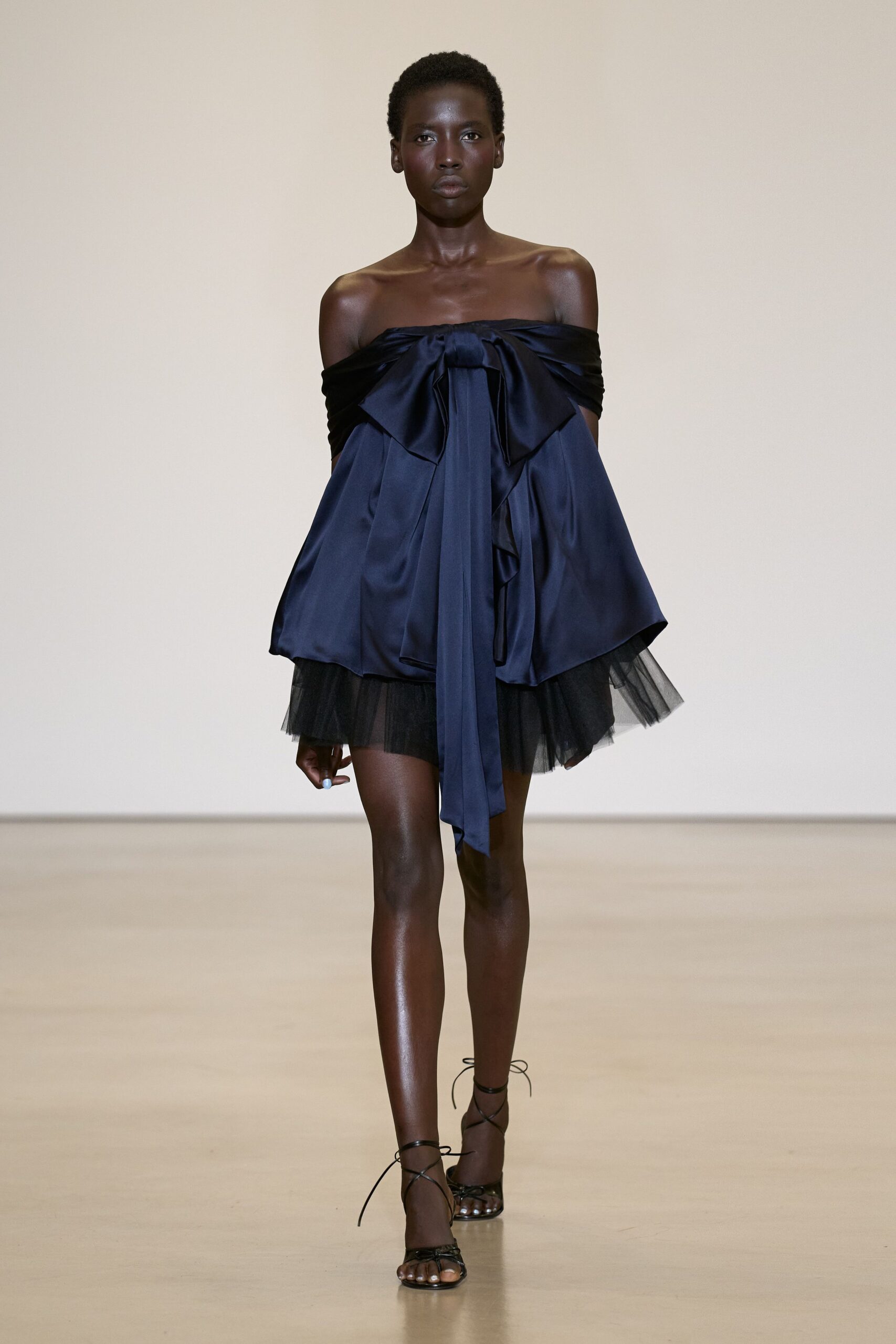 Midnight satin mini dress, off-the-shoulder fold tied in an oversized bow. Black tulle peeking below the hem in a soft uneven cloud. Black lace-up sandals with thin cords trailing past the ankles. For stepping onto a hotel terrace just after the band starts.