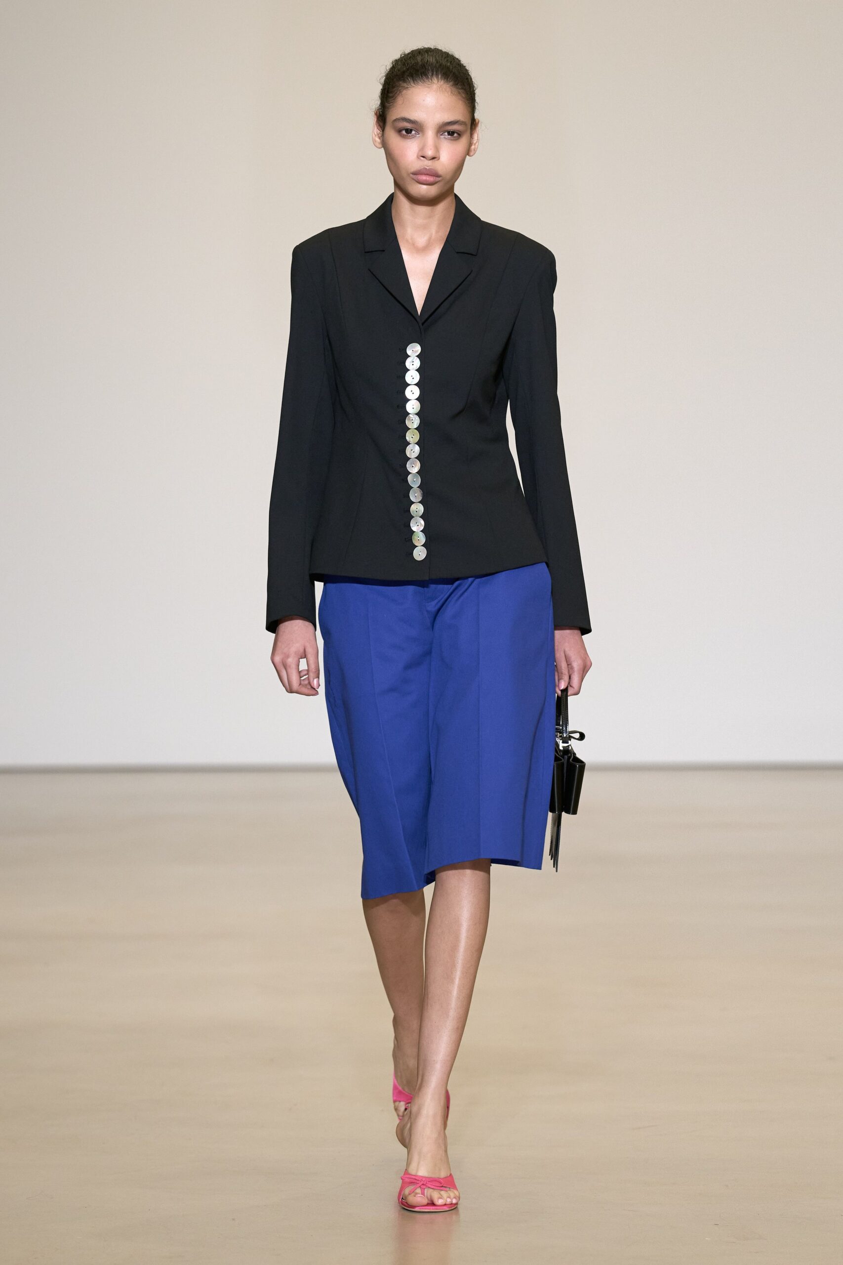 Black tailored blazer, a single column of pearly disc buttons off-center. Cobalt skirt cut on a slant, one hem corner dropping lower. Small black box bag with silver frame hardware. Hot-pink thong sandals with skinny knotted straps. Leaving a registry office with the wrong bouquet and better shoes.