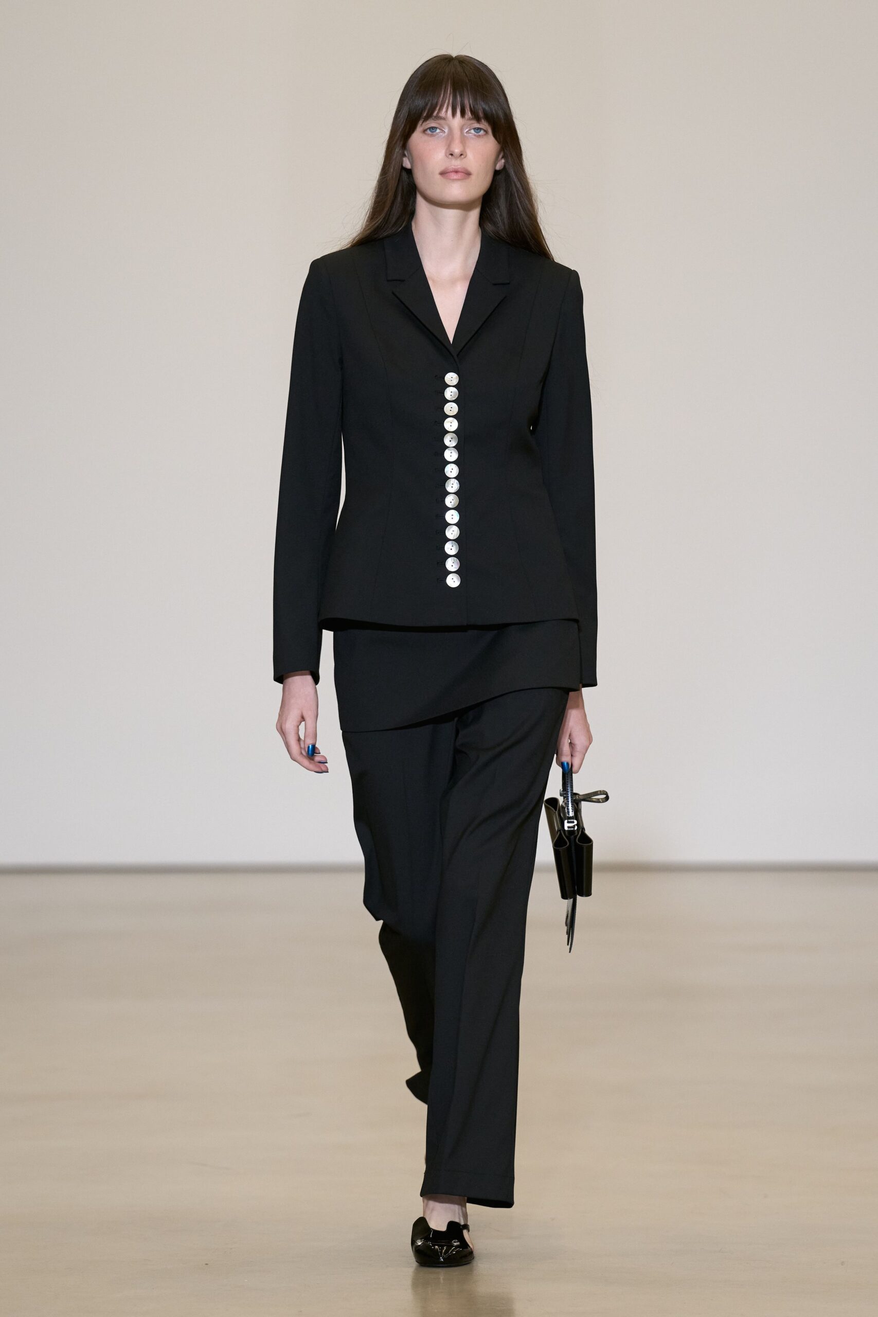 Black tailored blazer, a column of white buttons running off-center. Black straight trousers with a wrapped panel skimming one hip. Tiny black top-handle bag, folded into twin tubular pockets. Black patent loafers with silver bit hardware at the vamp. For signing paperwork in a hotel lobby before the rain starts.