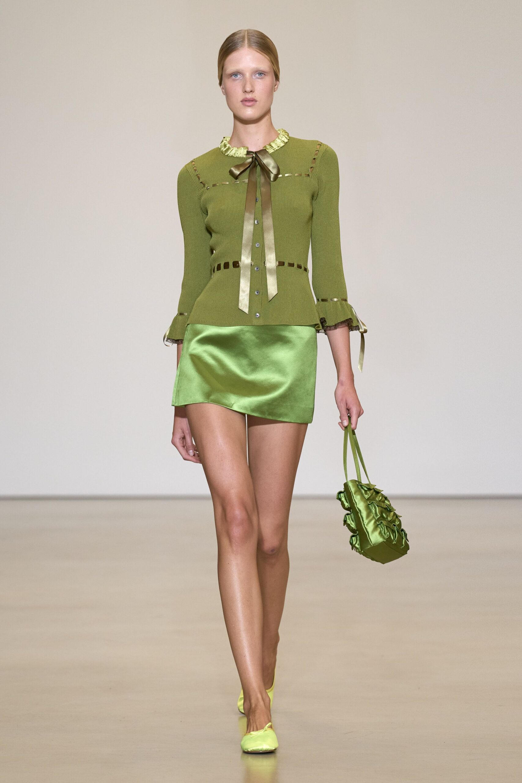 Moss ribbed cardigan with ribbon-threaded cutouts and a long satin bow. Acid-lime satin mini, cut on the bias with a soft ripple. Small satin shoulder bag puffed with ruffled side seams. Lime satin mules. Walking out of a florist with wet stems wrapped in brown paper.