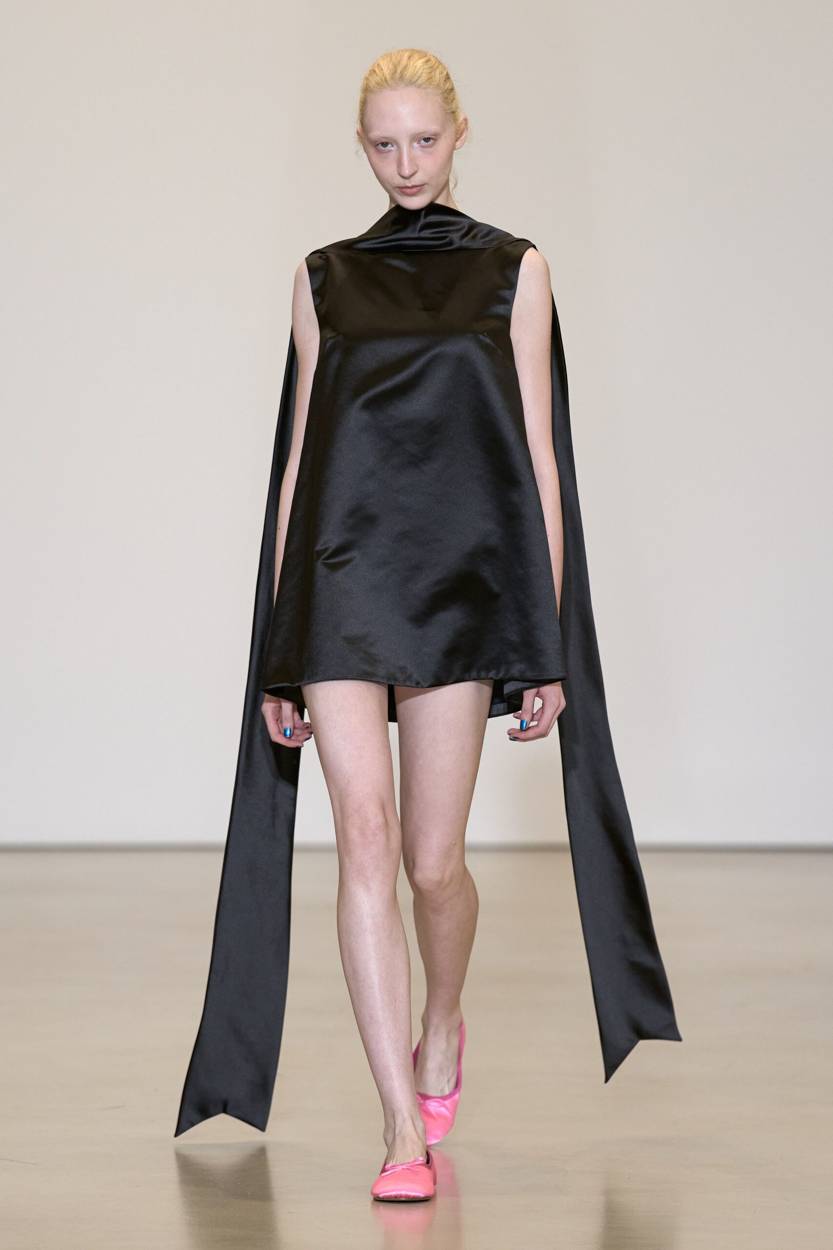 Black duchesse satin mini dress with a folded funnel neck and floor-sweeping side panels. Bubblegum satin flats with a soft collapsed square toe. For crossing a silent hotel lobby after midnight with your shoes still in hand.