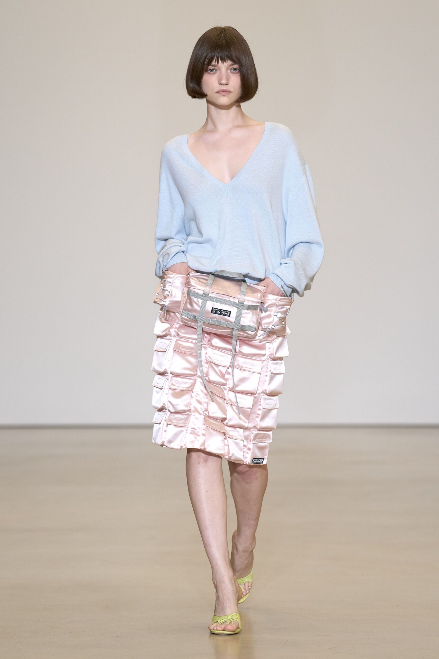 Powder-blue fine knit sweater with a deep V and dropped shoulders. Blush satin cargo skirt, stacked flap pockets and grey webbing cinched across the hips. Lime thong sandals with a tiny bow at the toe. Walking from a corner shop at dawn with flowers and yesterday's cash still stuffed away.