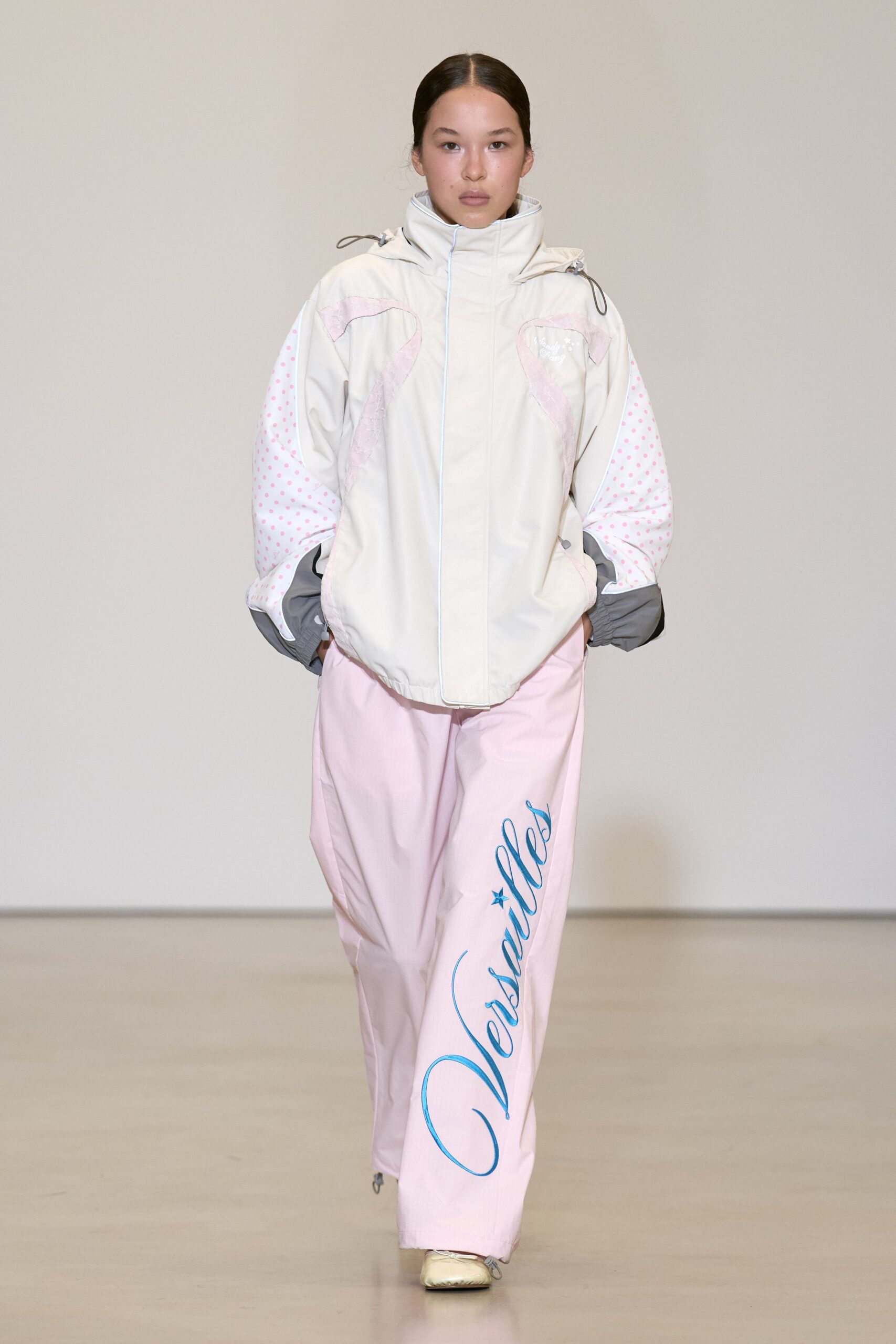Ivory nylon anorak with funnel neck, pink flocked swirls and dotted sleeves. Powder-pink ripstop track pants with oversized blue script curling down one leg. Cream satin ballet flats with thin tie laces. Walking the boardwalk early, before the amusement park opens.