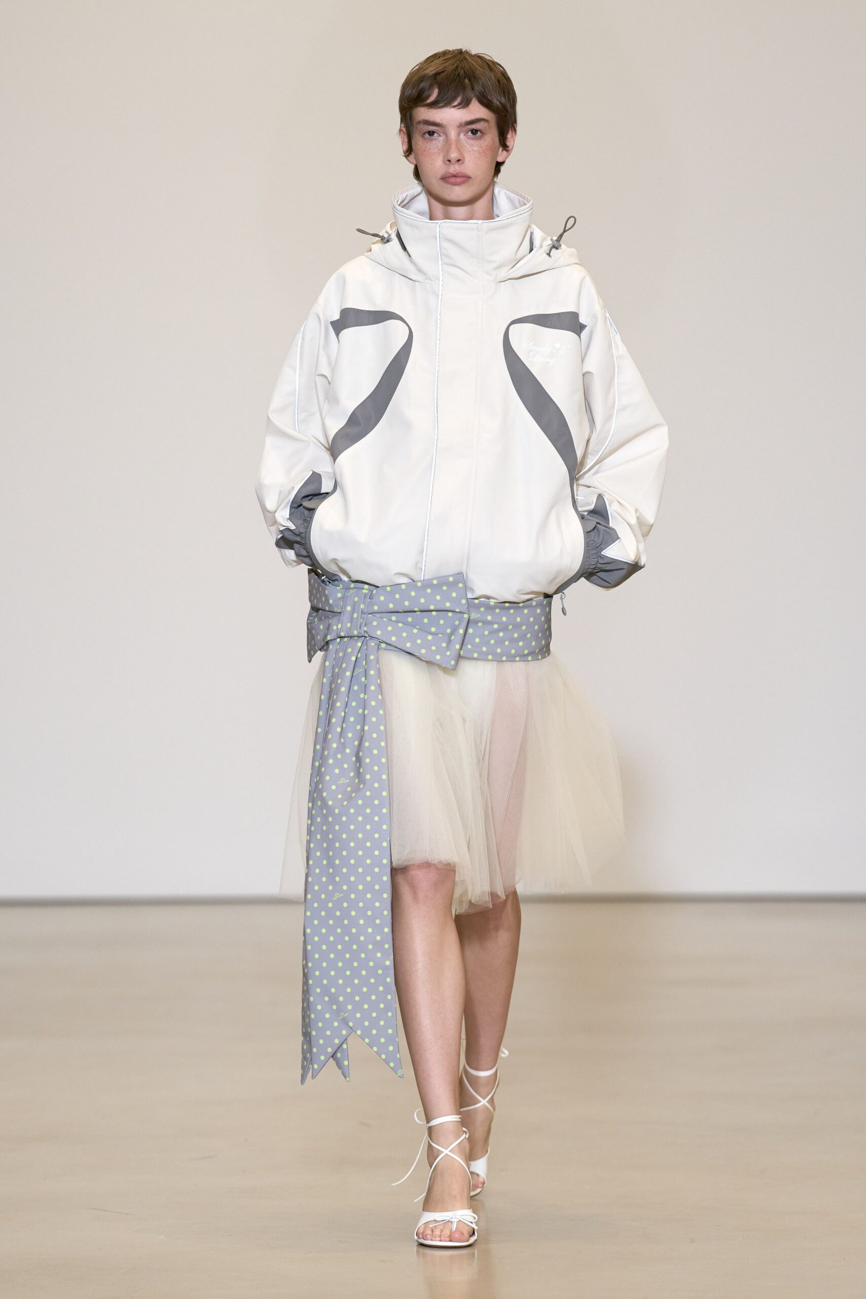 Ivory shell anorak with a tall funnel hood and grey swooping inserts. Wide grey sash belt dotted lime, one tail cut into swallow points. Cloudy ivory tulle skirt hanging sheer below the jacket hem. White lace-up sandals with thin cords crossing the instep. Crossing a windy pier with cake boxes balanced in both hands.